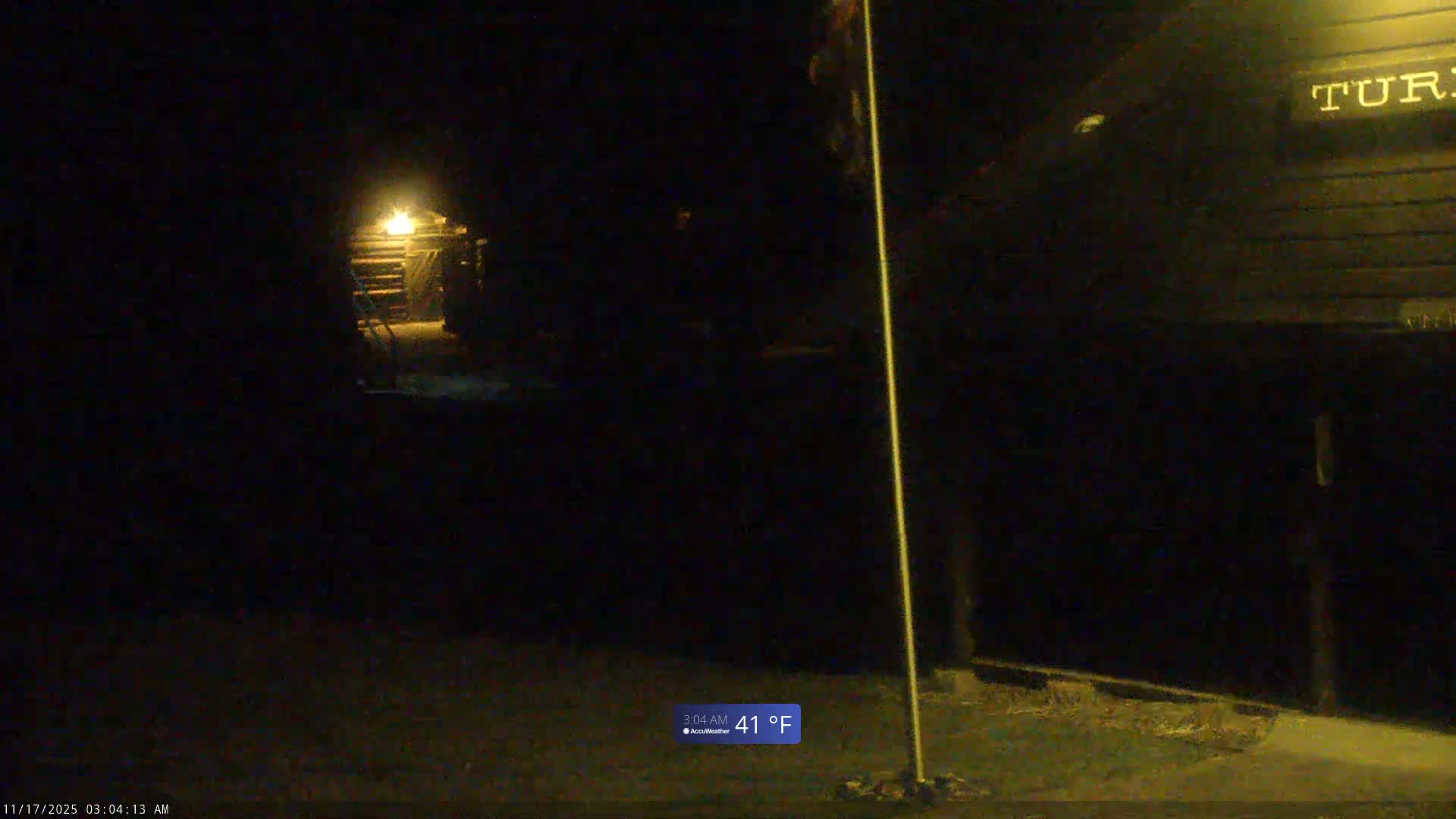 A dark outdoor scene at night, with a temperature of 41°F and clear conditions, shows a brightly lit building in the distance to the left, a tall flagpole in the foreground, and a shadowy building on the right.