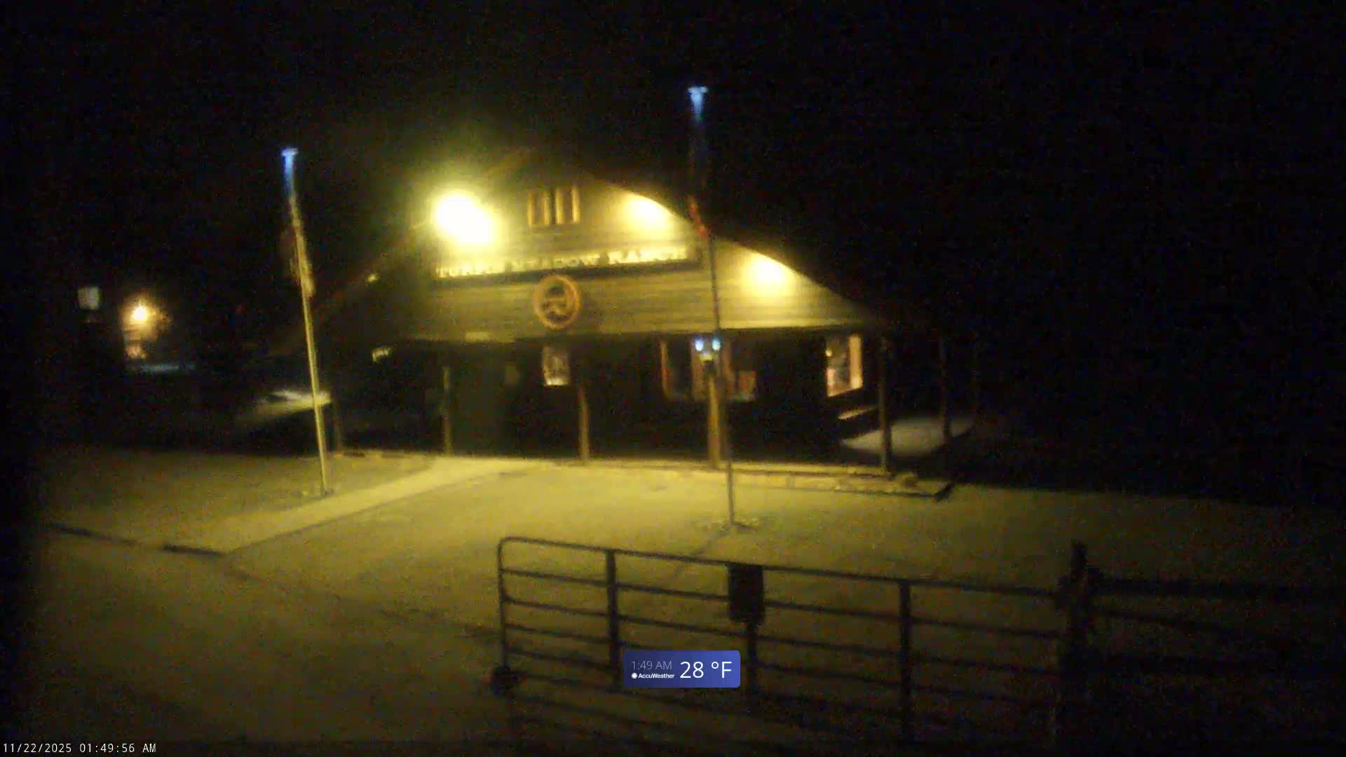 A rustic, illuminated lodge-style building with two flagpoles stands at night, seen behind a metal gate in the foreground, under clear conditions.