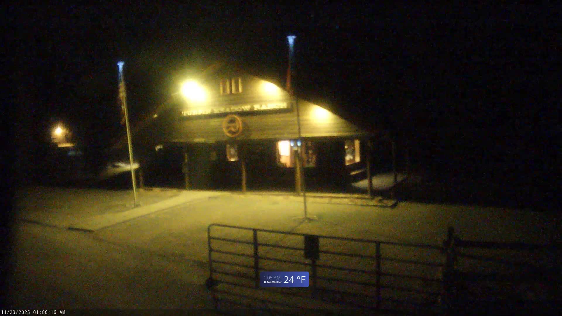 A warmly lit lodge or ranch building, featuring bright entrance lights and two flagpoles, stands on a paved area with a dark metal fence in the foreground, under a clear, dark 24°F night.