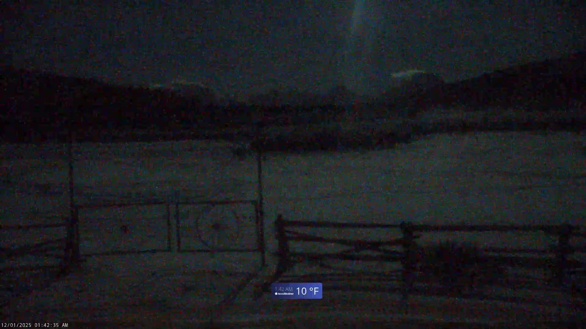 A dark, grainy image captures a snow-covered outdoor landscape at night, featuring a fence in the foreground and dark, distant hills under a hazy sky, indicating cold winter weather.