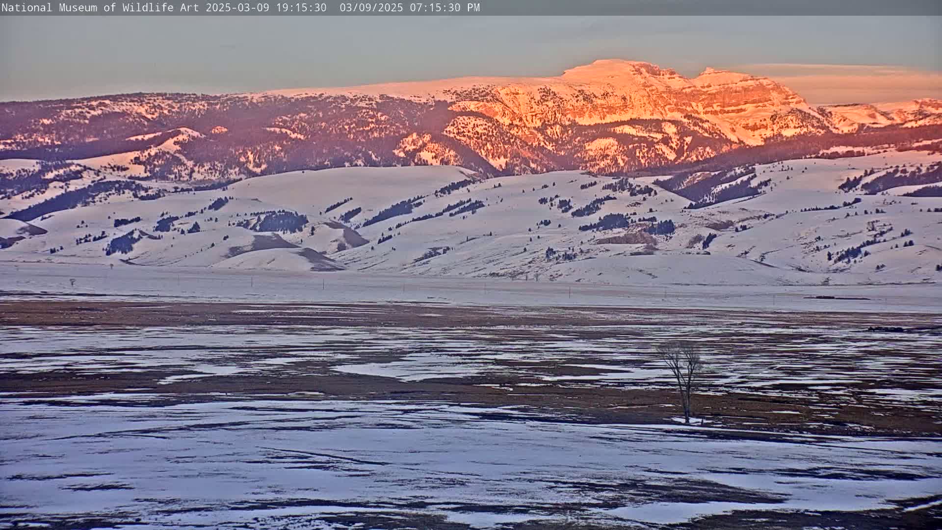 A snow-covered landscape with a mountain range in the background illuminated by the setting sun.