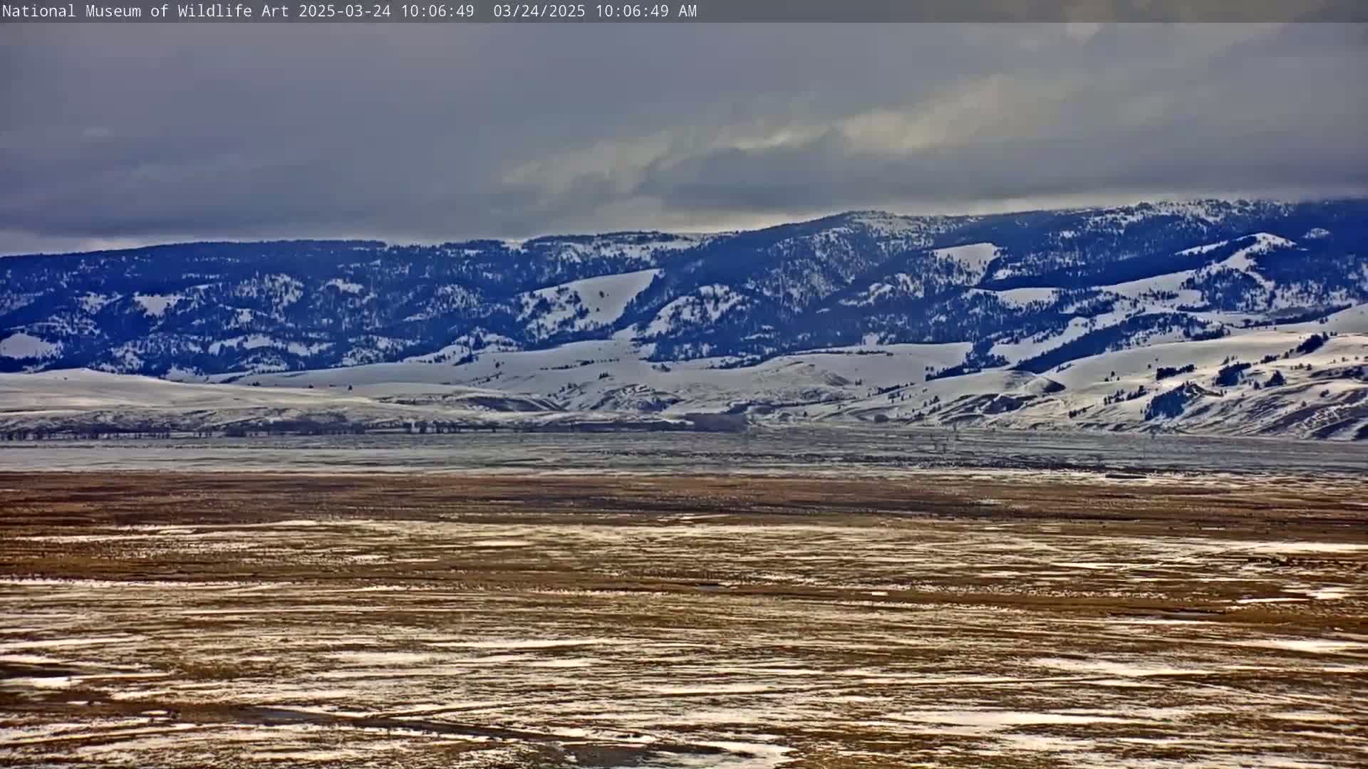 A snow-covered mountain range is visible in the distance under a cloudy sky, with a flat, mostly brown expanse of land speckled with snow in the foreground.