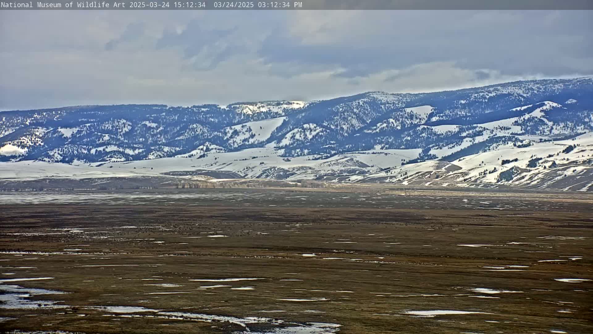 A snow-dusted mountain range is visible in the distance behind a wide, flat plain with patches of snow and ice under a mostly cloudy sky.