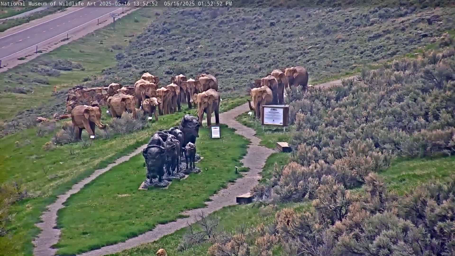 A herd of elephant and bison sculptures stand on a grassy hillside overlooking a highway, under a partly cloudy sky.