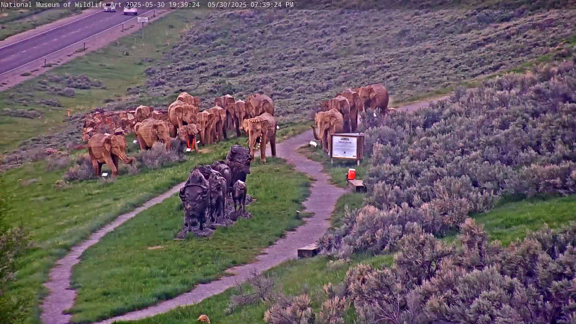 A grassy hillside with numerous elephant and bison sculptures, a paved pathway, and a highway visible in the distance under a clear evening sky.