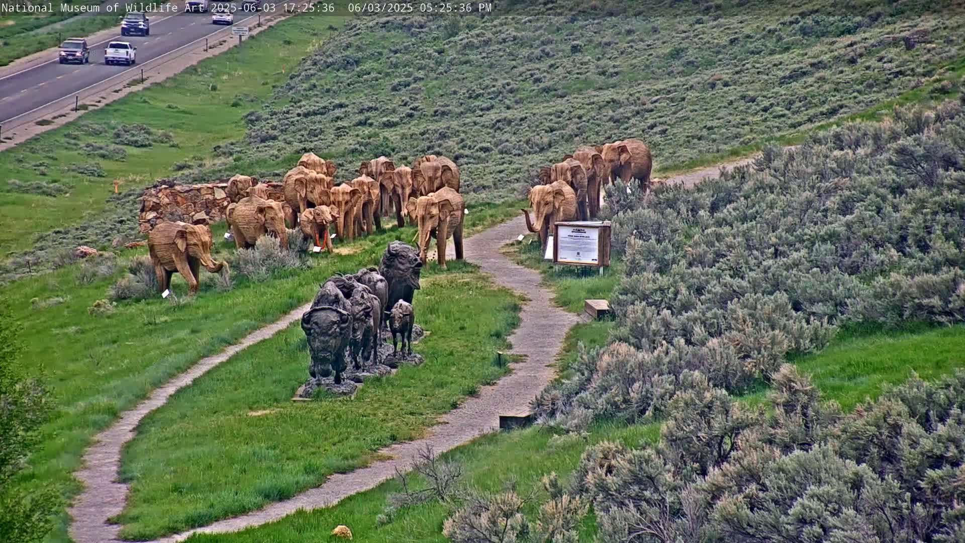 A winding path leads through a grassy area with sculptures of elephants and bison, overlooking a highway with cars.