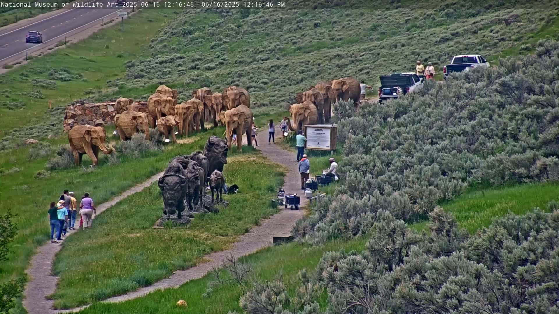 A winding path leads to several large sculptures of elephants and bison, with people observing them on a grassy hillside under a sunny sky.