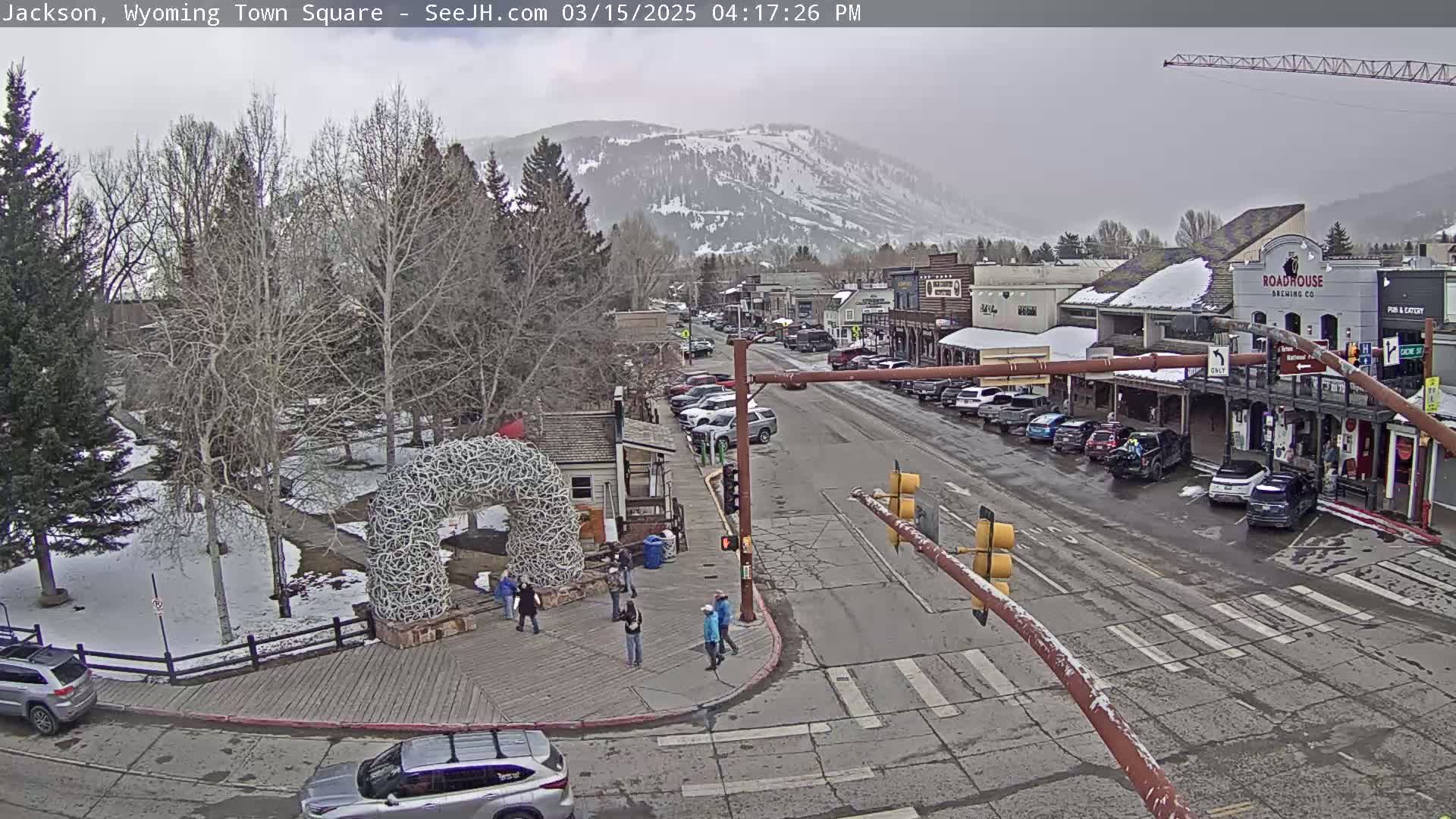 A snow-dusted town square scene shows a main street with parked cars, pedestrians near an antler arch, and a snow-covered mountain in the background under an overcast sky.