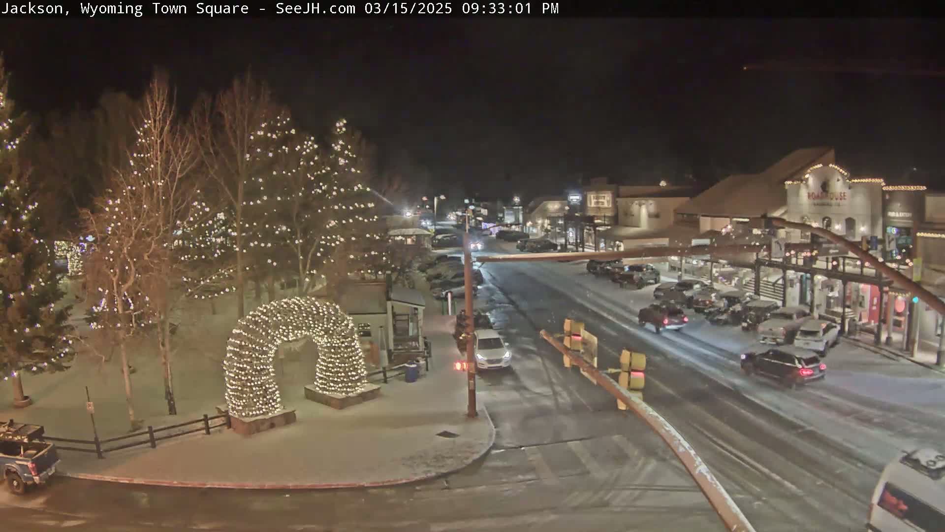 A snow-covered town square at night, illuminated by string lights on trees and an archway, with cars driving on a street lined with buildings.