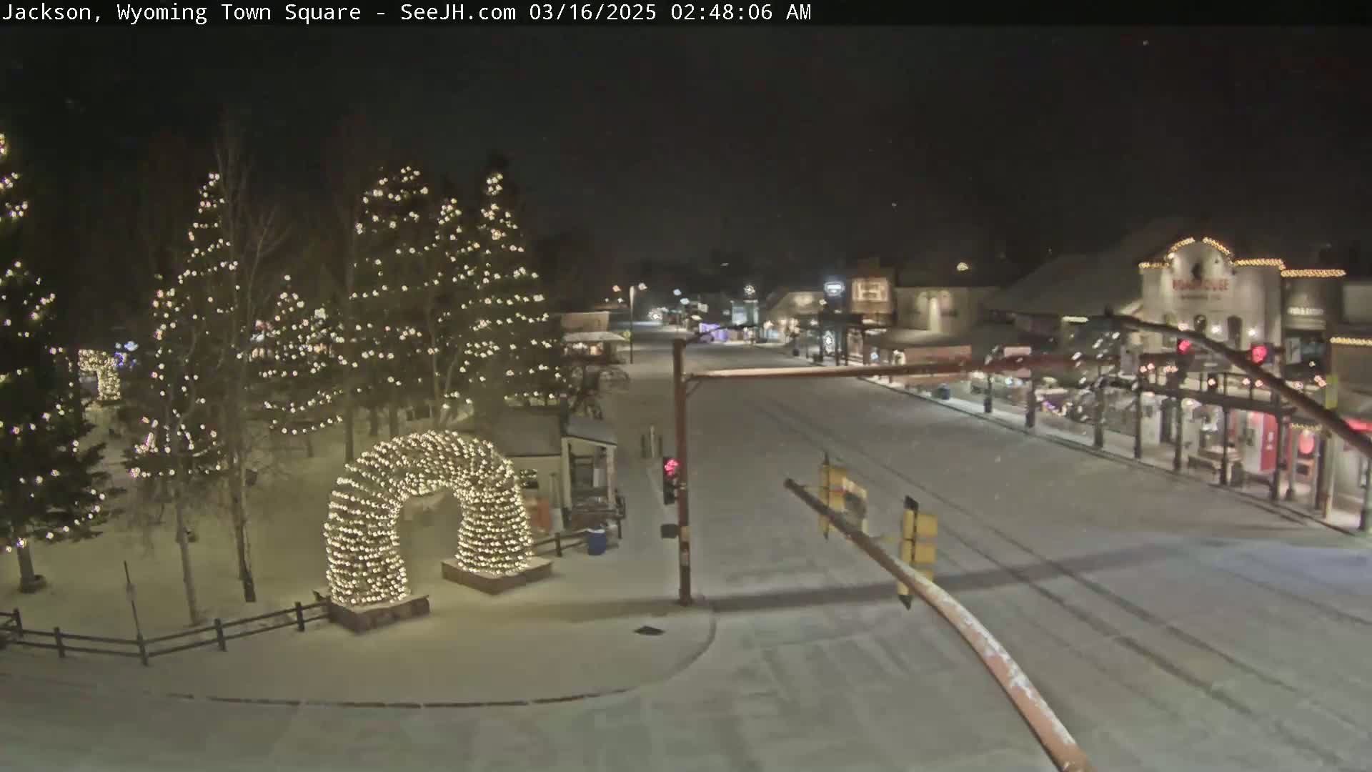 A snow-covered town square at night, illuminated by Christmas lights on trees and an archway, with a quiet street visible in the background.