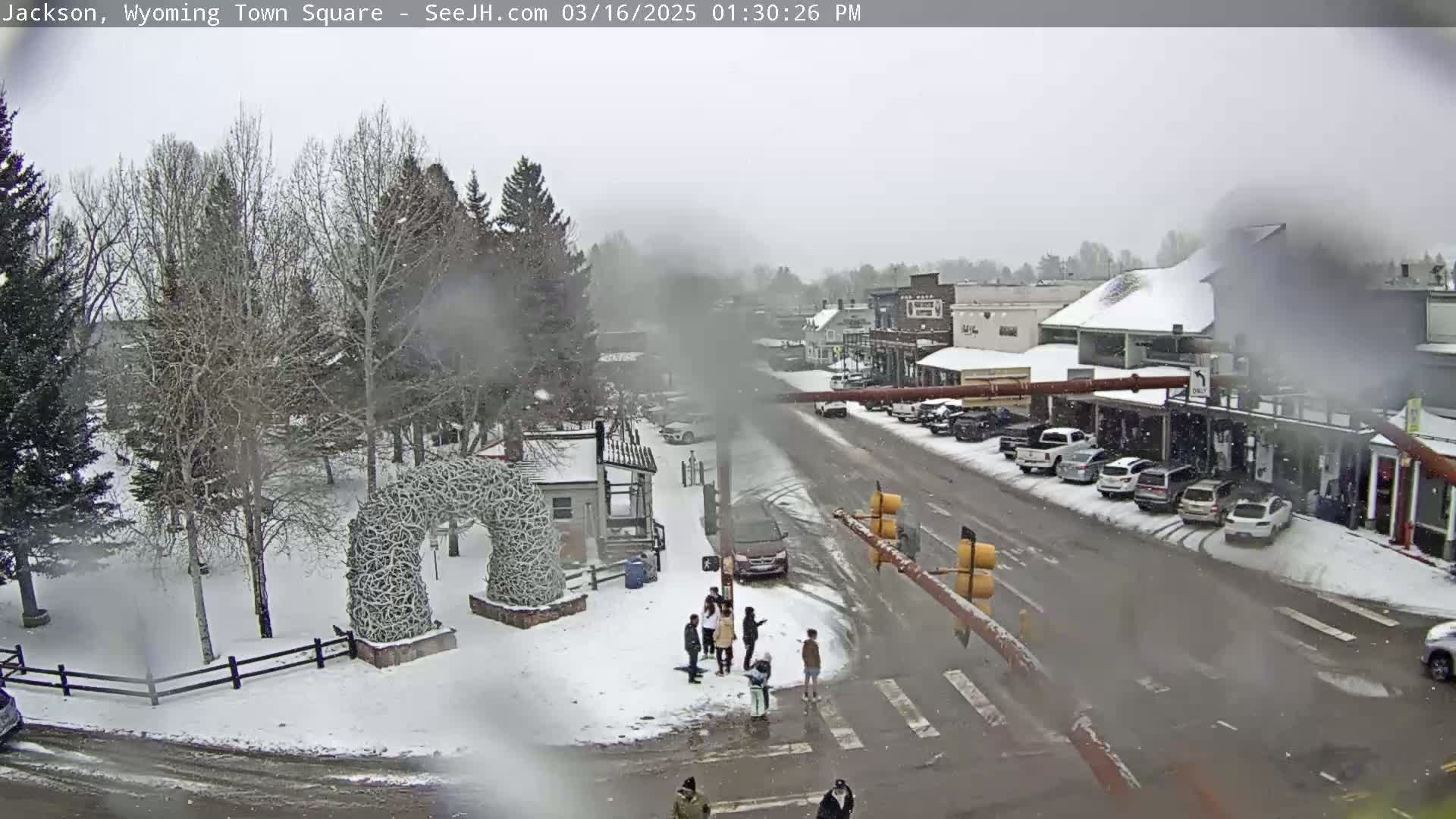 A snow-covered town square with a few pedestrians and parked cars, under a lightly snowing sky.