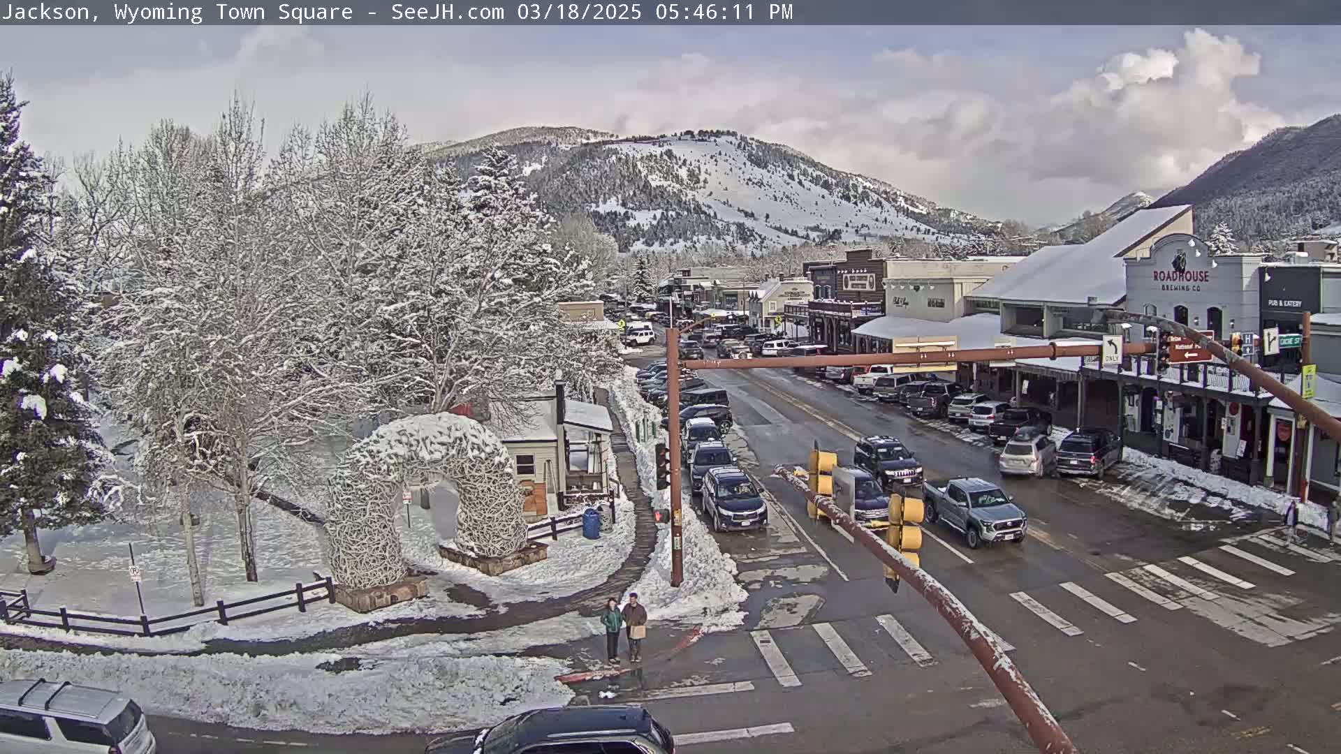 A snow-covered town square with cars parked along the street, pedestrians crossing, and snow-covered trees and mountains in the background under a partly cloudy sky.