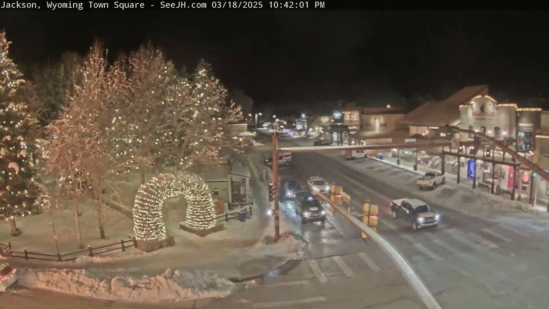 A snow-covered town square at night, illuminated by lights on trees and a large arch, with cars driving on a nearby street.