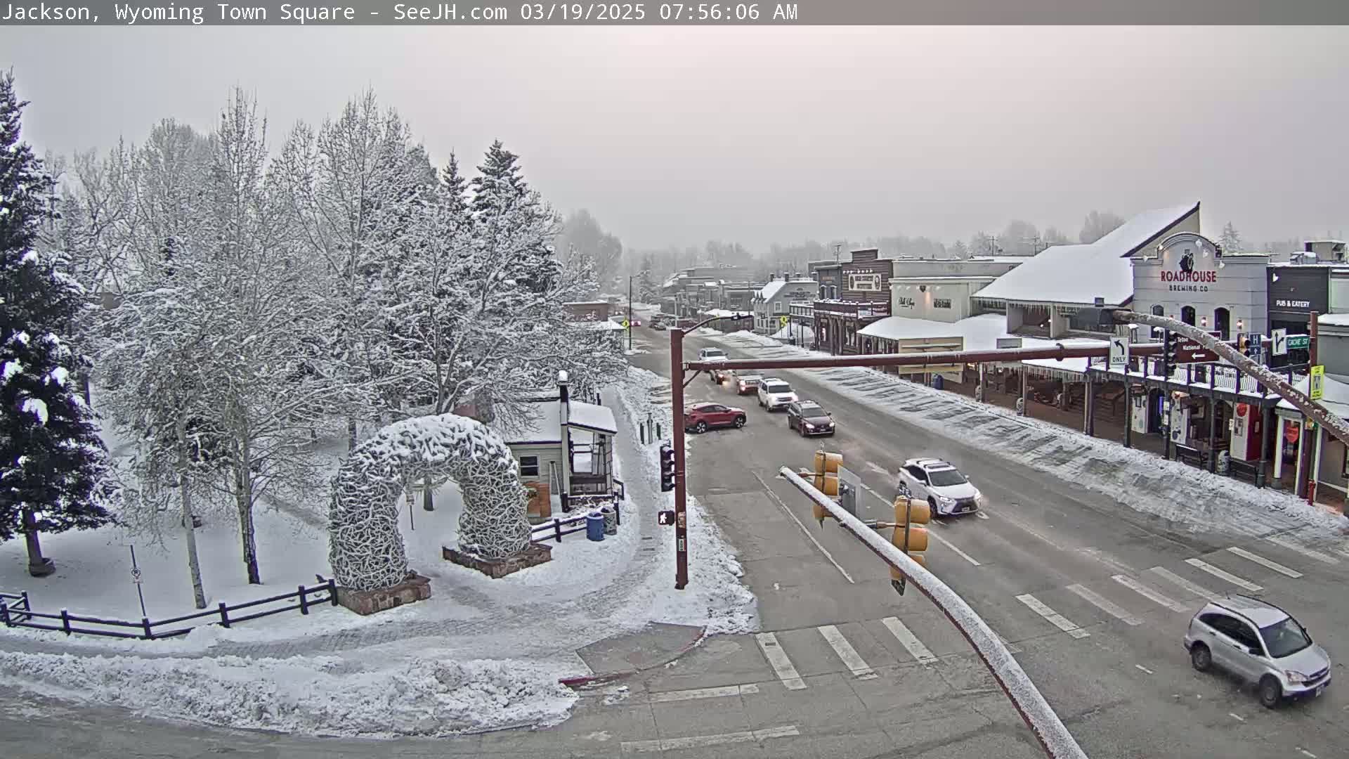 A snow-covered town square with several cars driving on a street, surrounded by snow-laden trees and buildings.