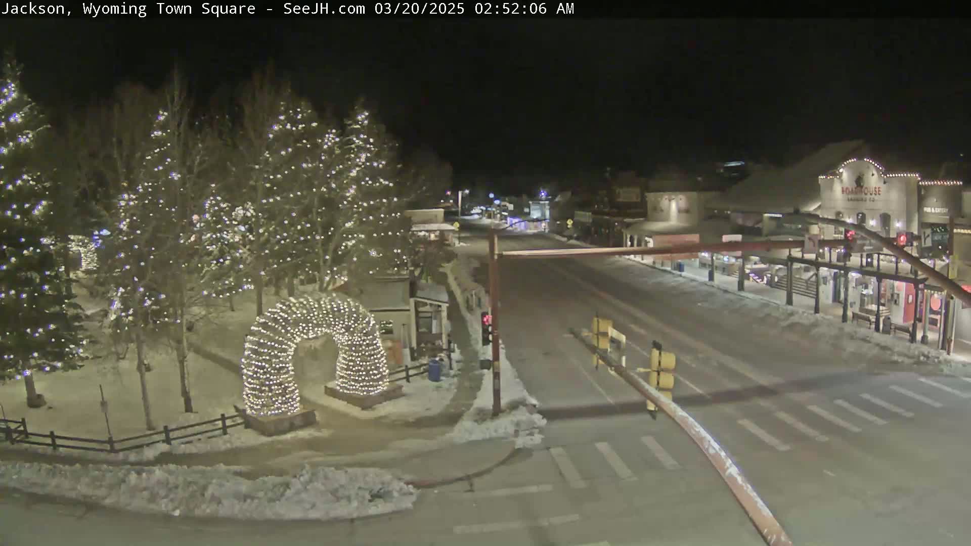 A snow-covered town square at night, illuminated by strings of lights on trees and an archway, with a quiet street visible beyond.