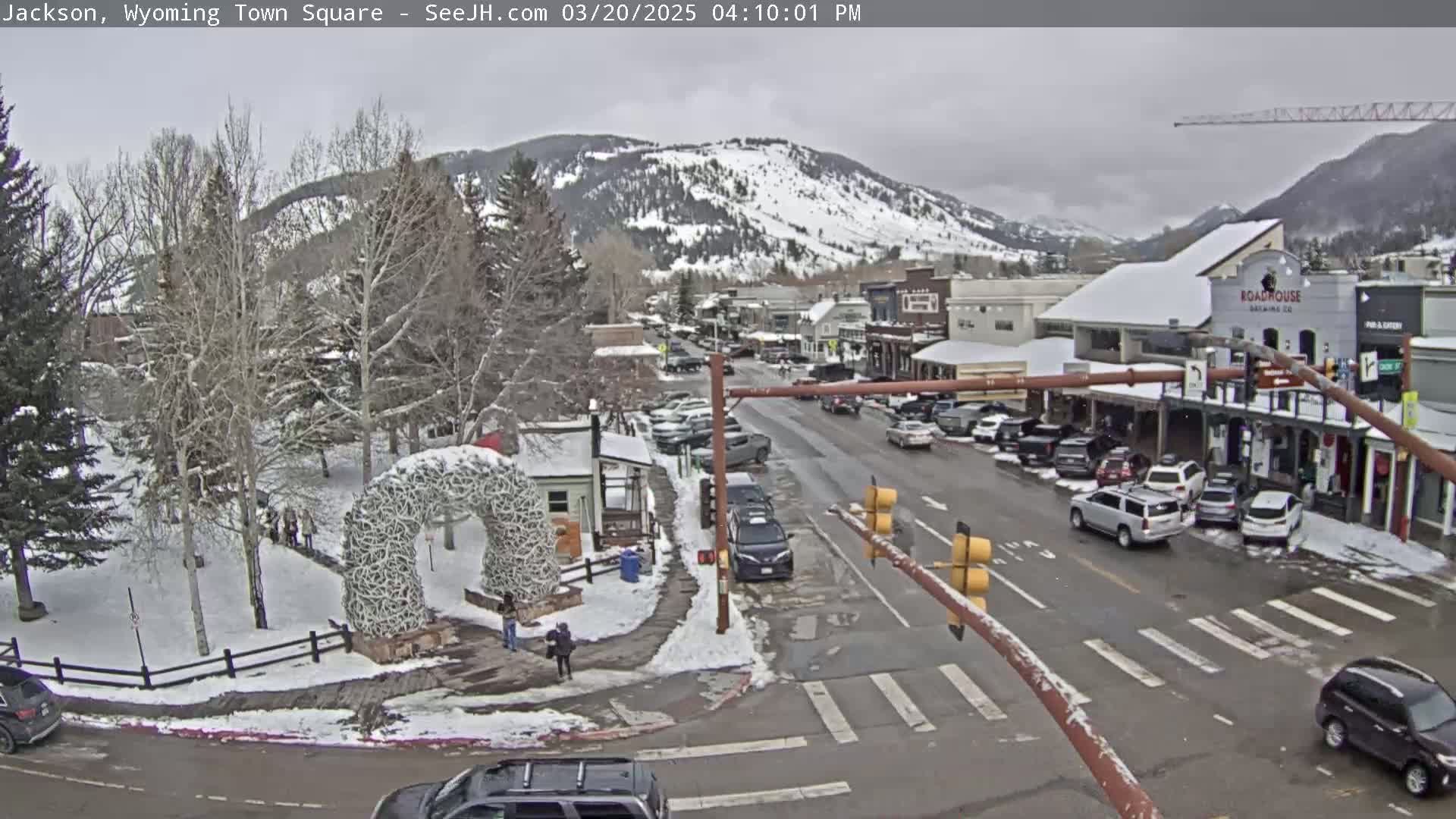 A snow-covered town square with cars parked along the street, a large antler arch in a park, and snow-capped mountains in the background under an overcast sky.