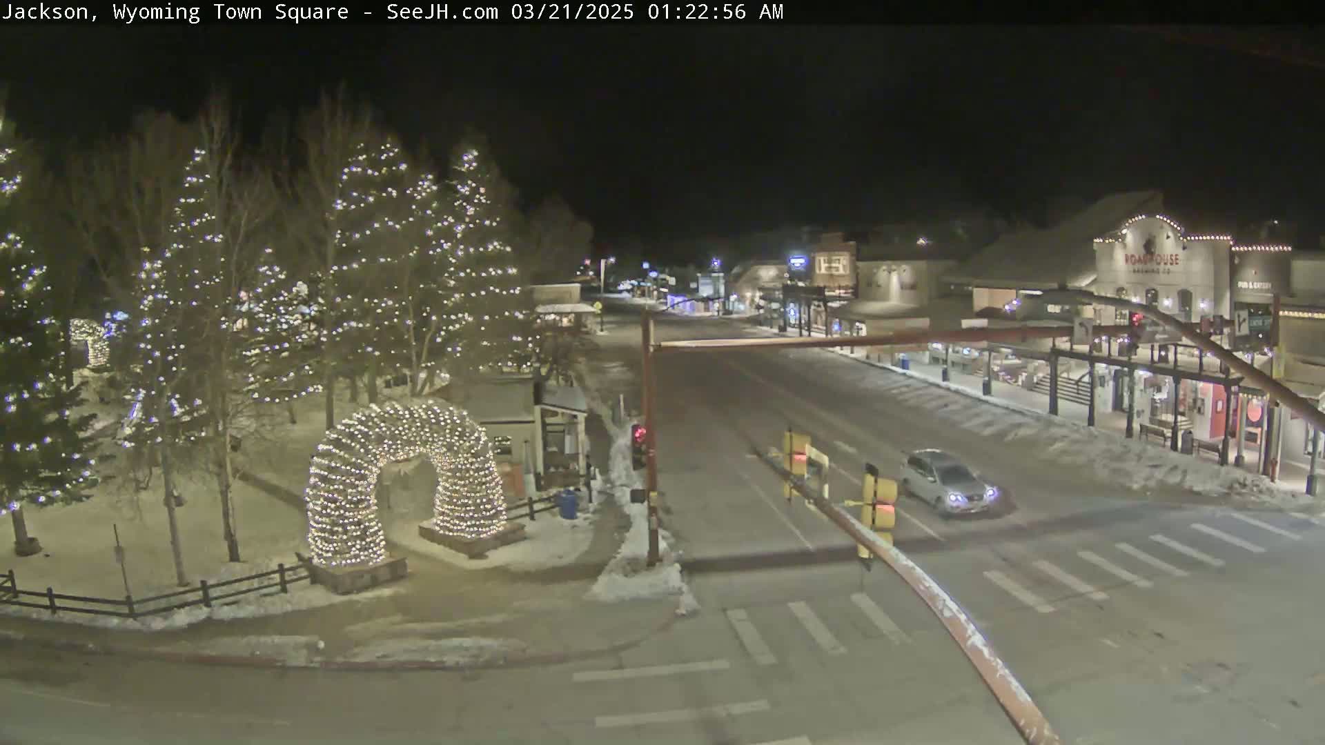 A snow-covered town square at night, illuminated by Christmas lights on trees and an archway, with a single car driving on a road lined with buildings.