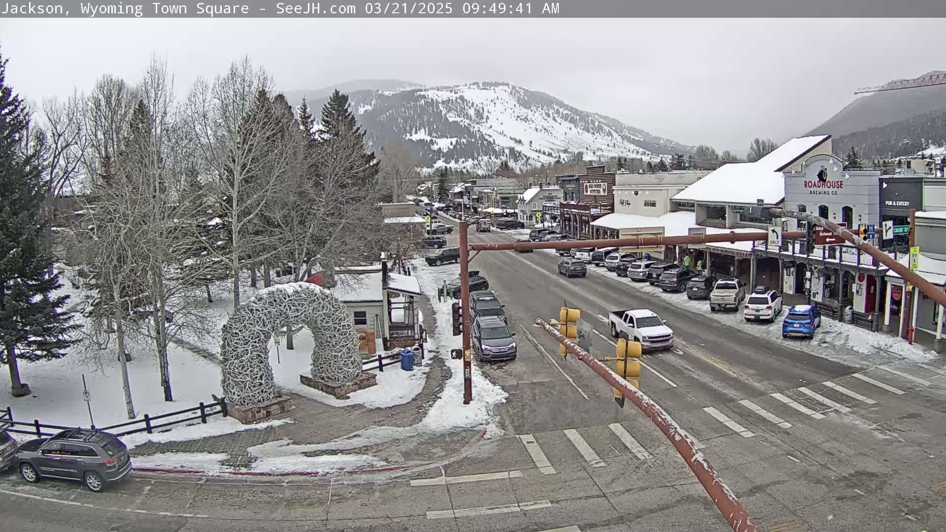 A snow-covered town square with parked cars and buildings lines a street, under an overcast sky, with a snow-capped mountain in the background.