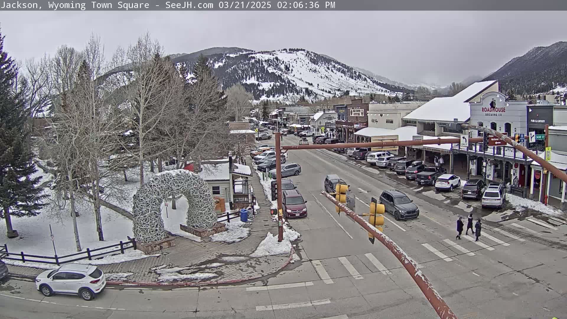 A snow-covered town square with cars parked along the street and a few pedestrians crossing, situated at the base of a snow-capped mountain range under a cloudy sky.