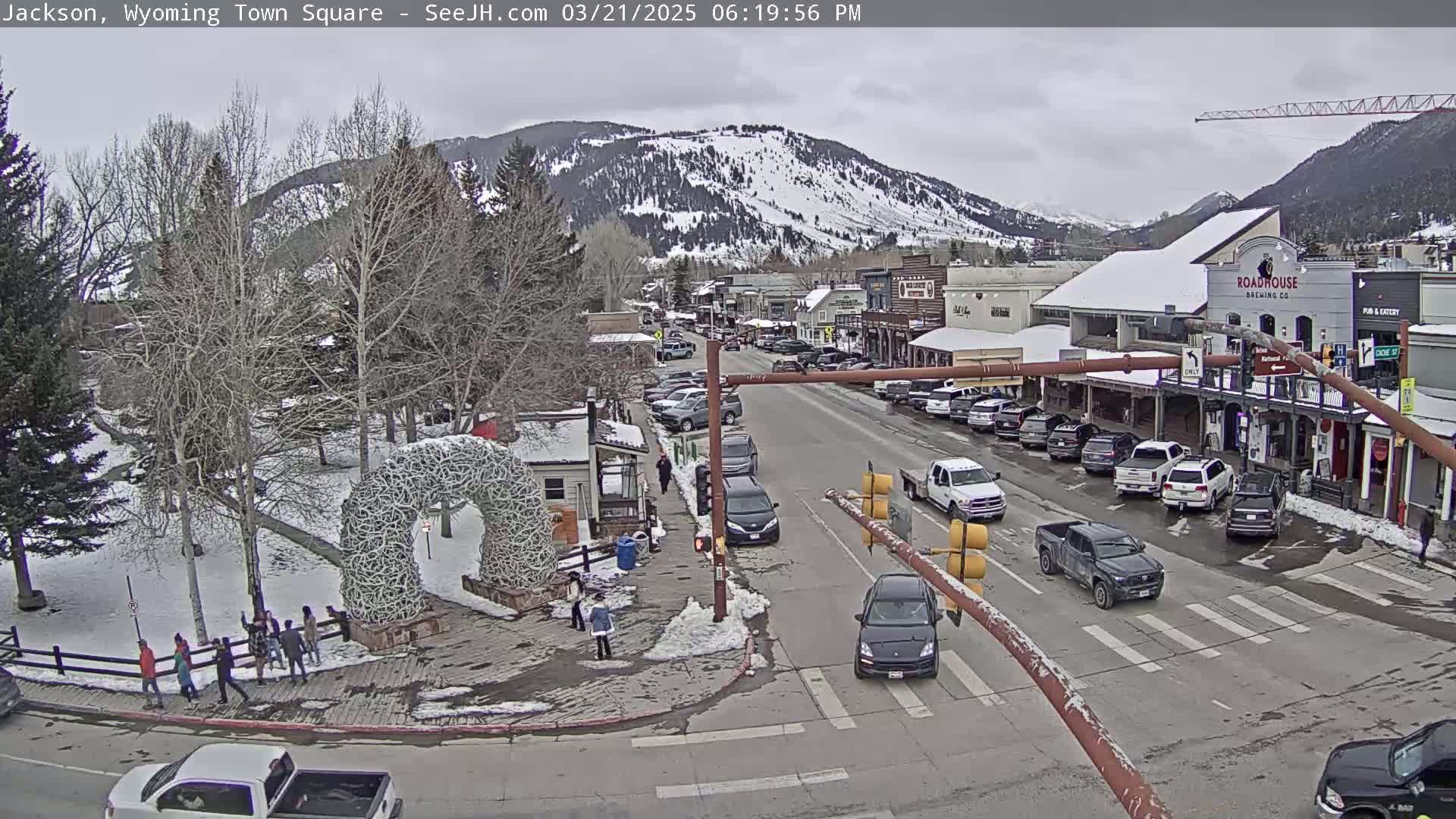 A snow-dusted town square, nestled in a valley beneath snow-capped mountains, shows a street scene with parked and moving cars under an overcast sky.