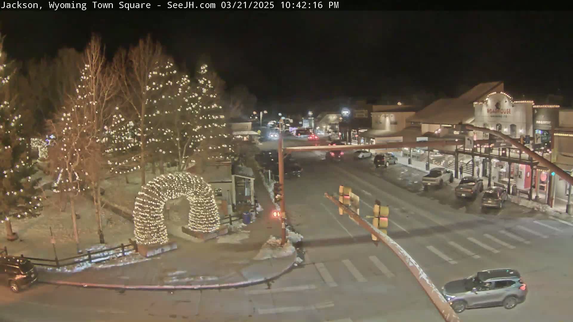 A nighttime view of a town square, decorated with Christmas lights, shows lightly snow-covered ground and cars driving on the street.