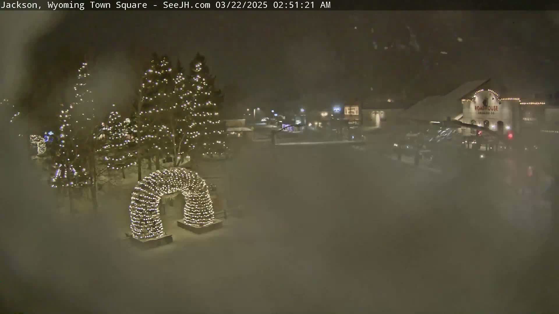 A snow-covered town square at night, illuminated by Christmas lights strung on trees and an archway, with a hazy, snowy atmosphere.