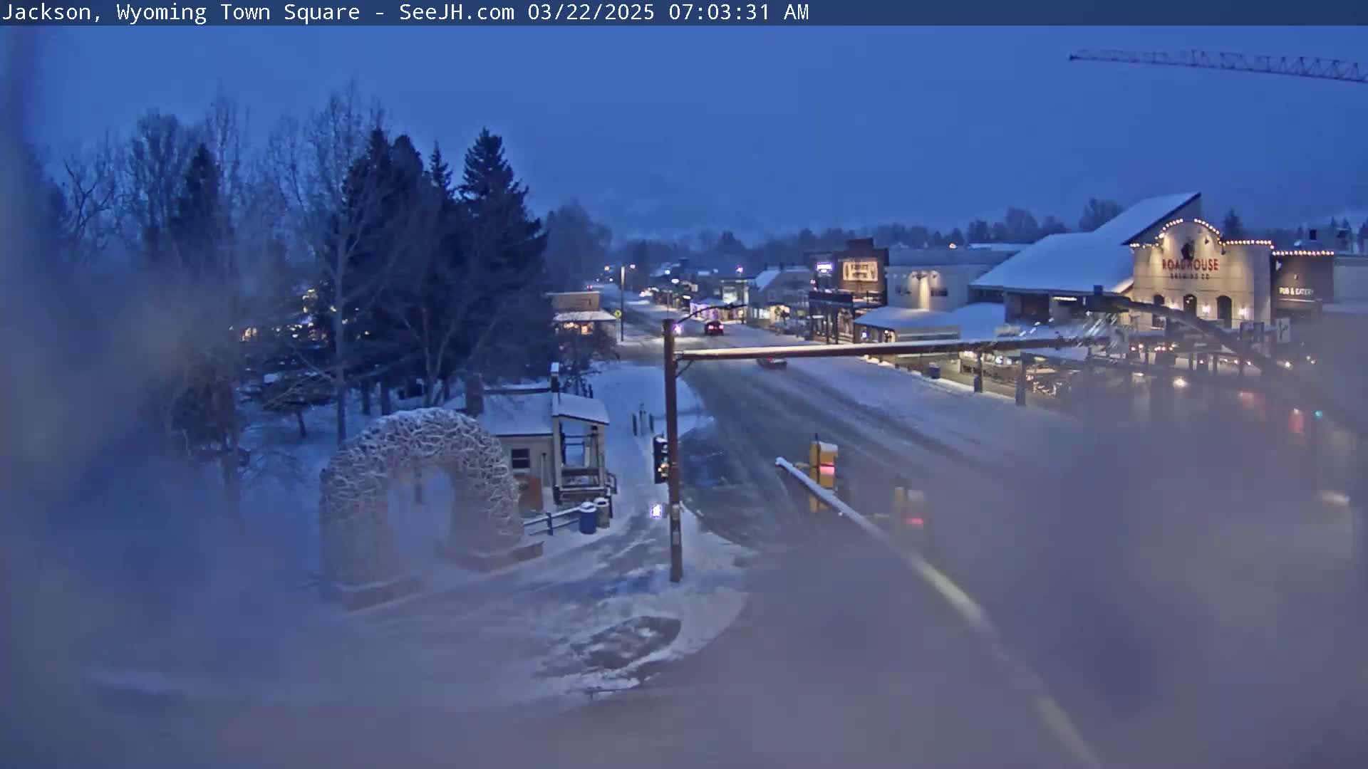A snow-covered town square at twilight, with a few cars on the road and snow-covered trees and buildings in the background.