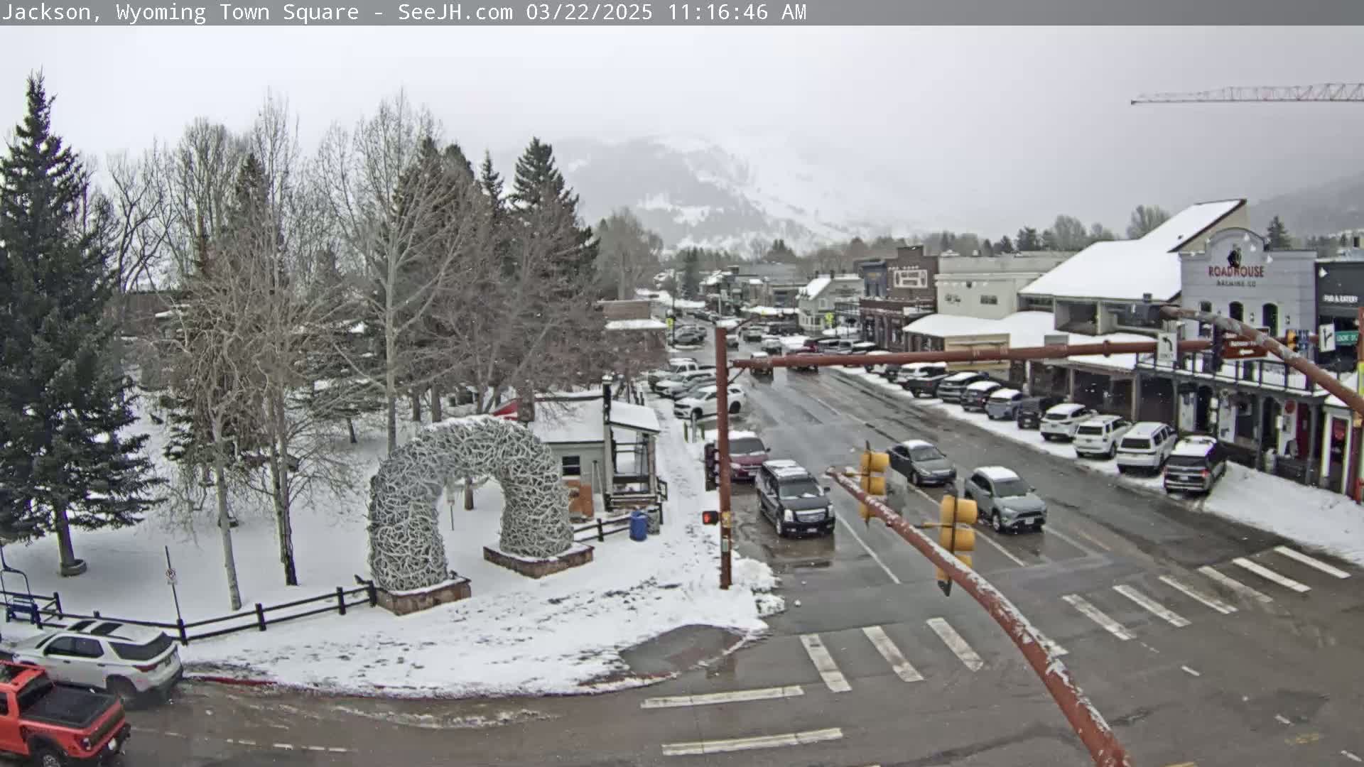 A snow-covered town square with cars parked along the street and a large antler arch in the foreground, under a lightly snowing sky with mountains in the background.