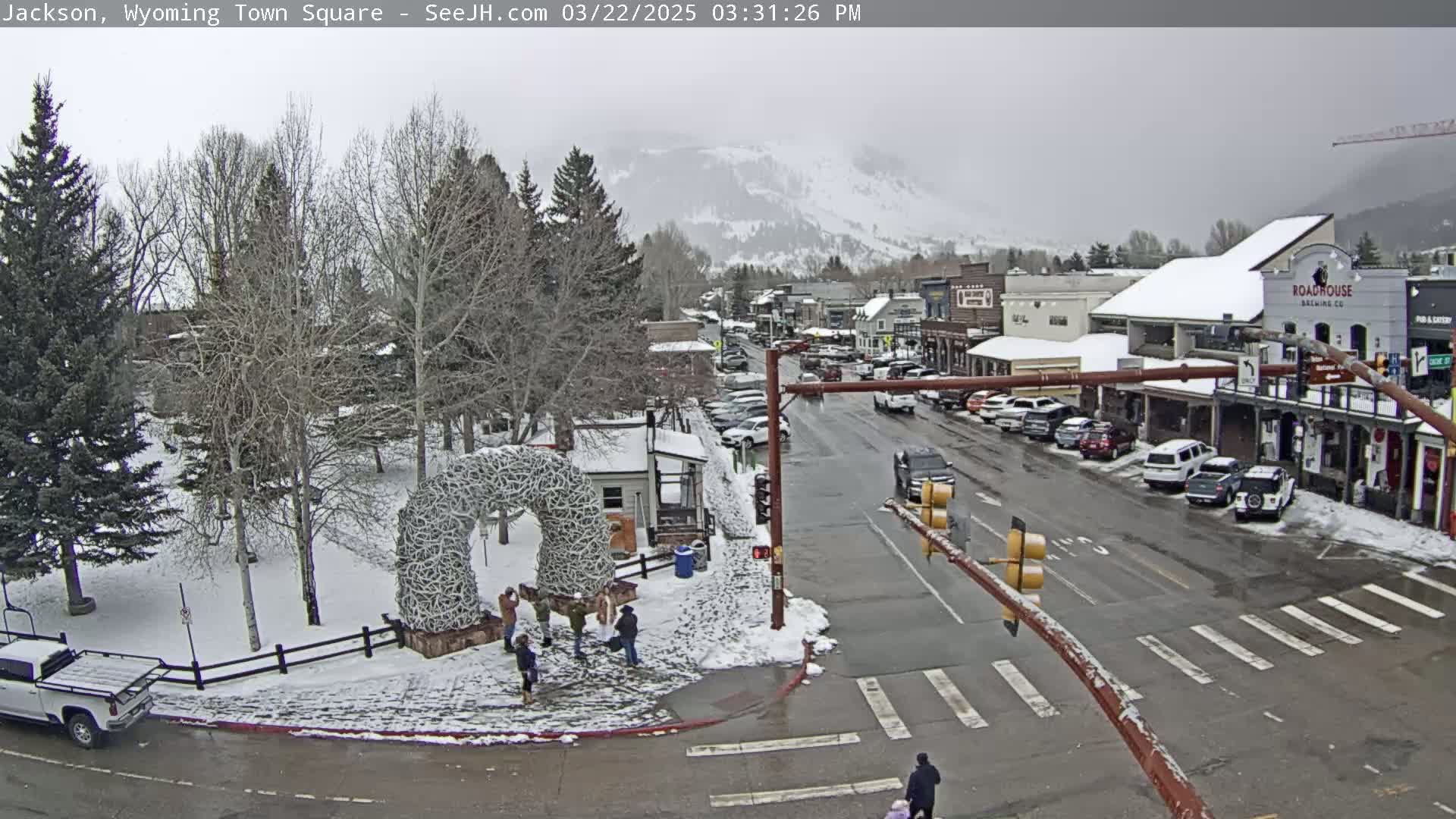 A snow-covered town square with a large antler arch, several parked cars lining the street, and a few pedestrians are visible under an overcast sky.