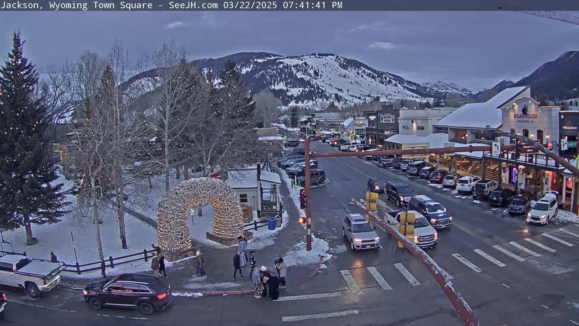 A snow-dusted town square at dusk, with cars on the street and pedestrians near a light-covered archway, is nestled against a mountain backdrop.