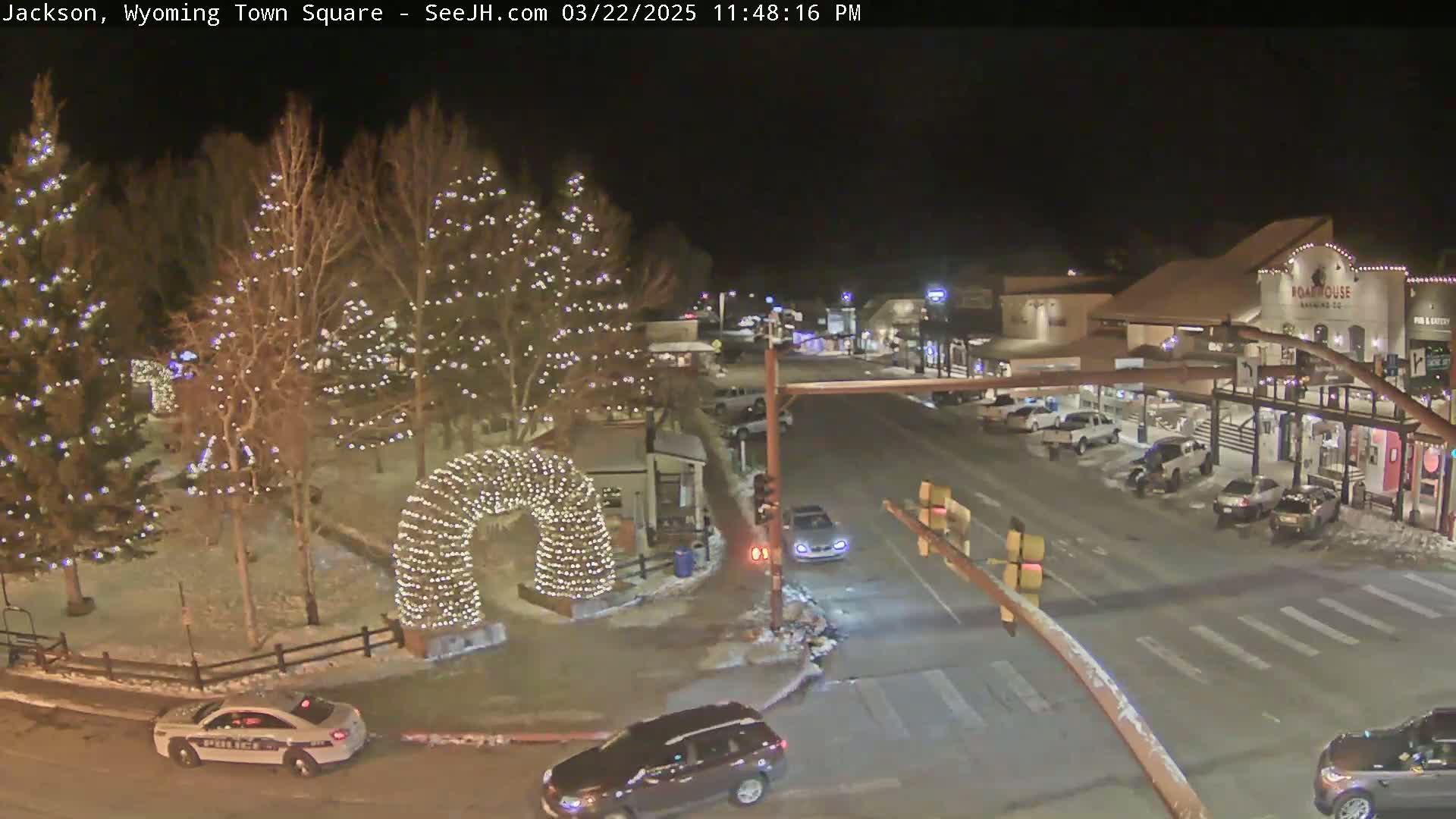 A nighttime view of a town square decorated with Christmas lights, featuring snow-covered ground, parked cars, and a police car.