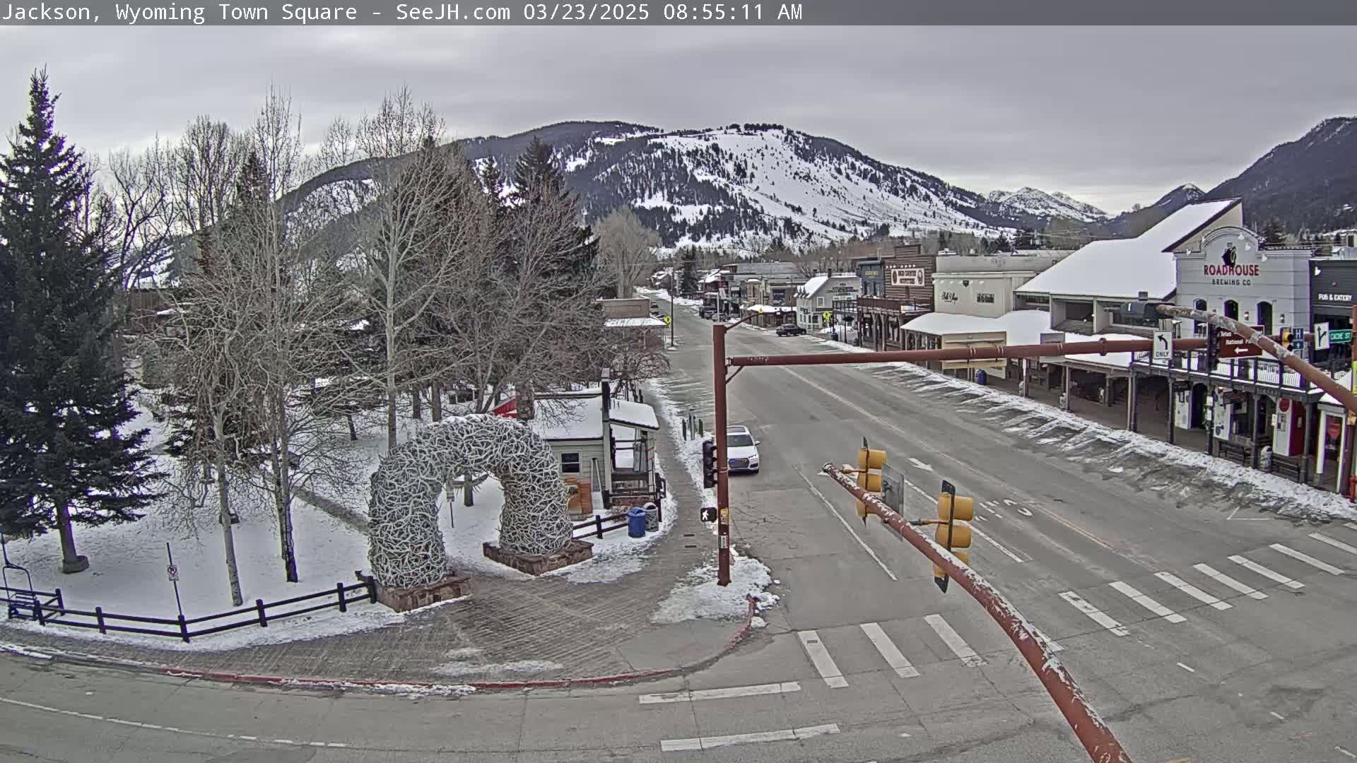 A snow-covered town square and street scene are visible under an overcast sky, with mountains in the background showing some patches of snow.