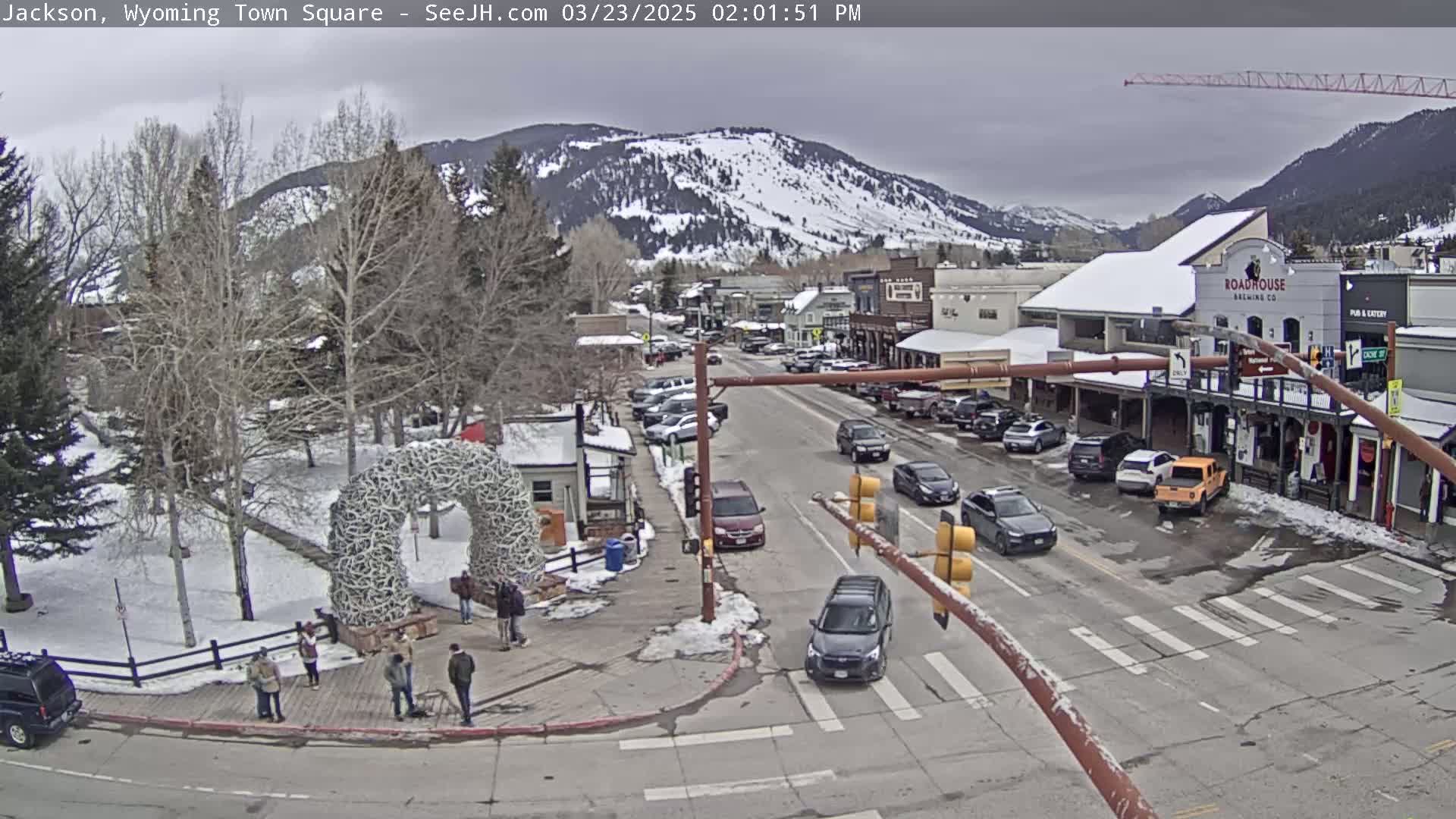 A town square with a large antler arch, surrounded by buildings and parked cars, under an overcast sky with snow-covered mountains in the background.