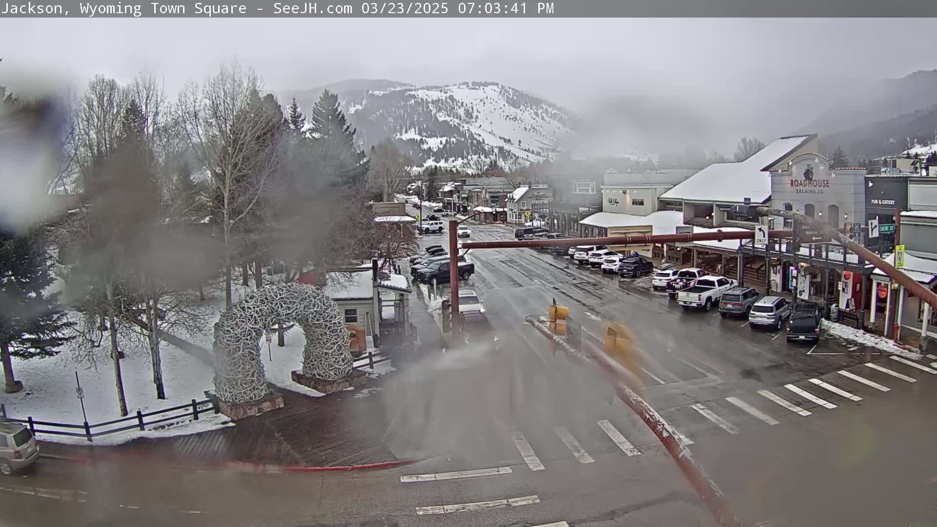 A snow-dusted town square with cars parked along the sides of a street, under an overcast sky.