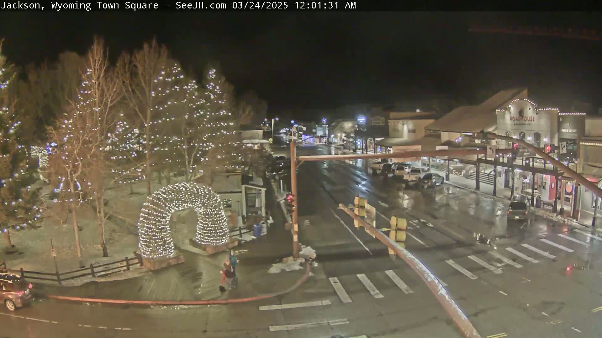A town square at night, lightly snowing, with Christmas lights decorating trees and an archway.
