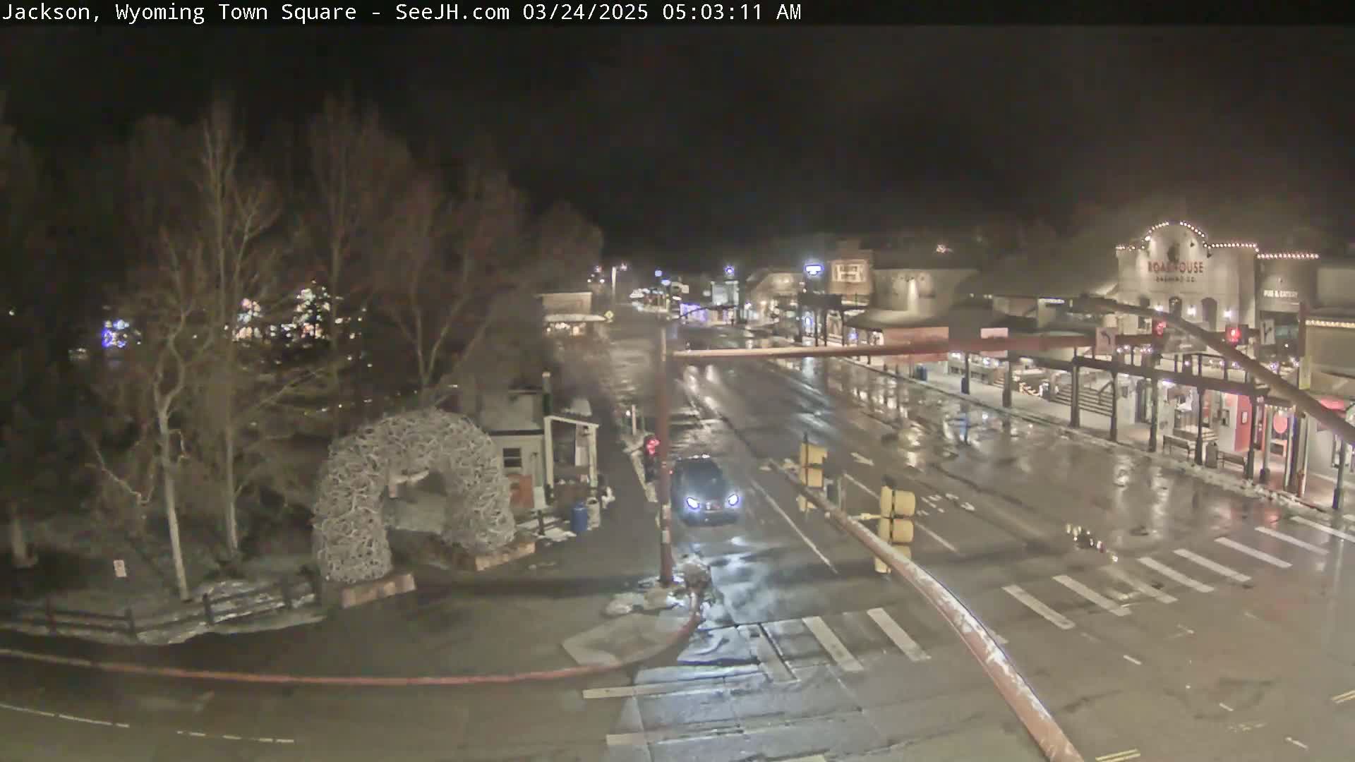 A wet, nighttime view of a town square with sparse traffic and bare trees under dim lighting.