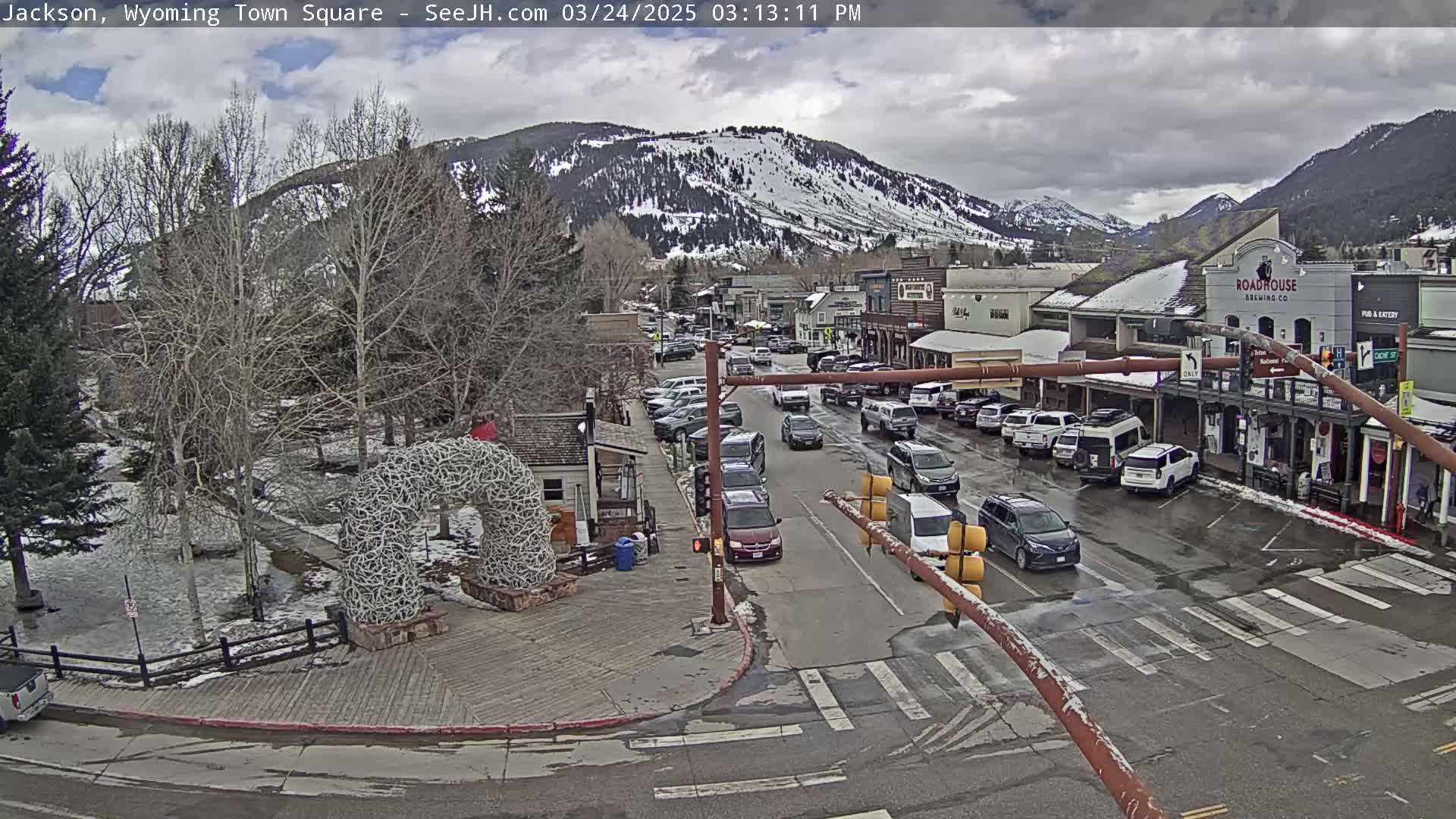 A town square with parked cars and traffic, a large antler arch, and snow-capped mountains in the background under a partly cloudy sky.