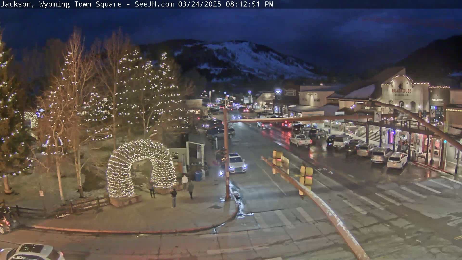 A town square at night, illuminated by Christmas lights on trees and an archway, shows a street with cars and a snowy mountain in the background under a dark sky.
