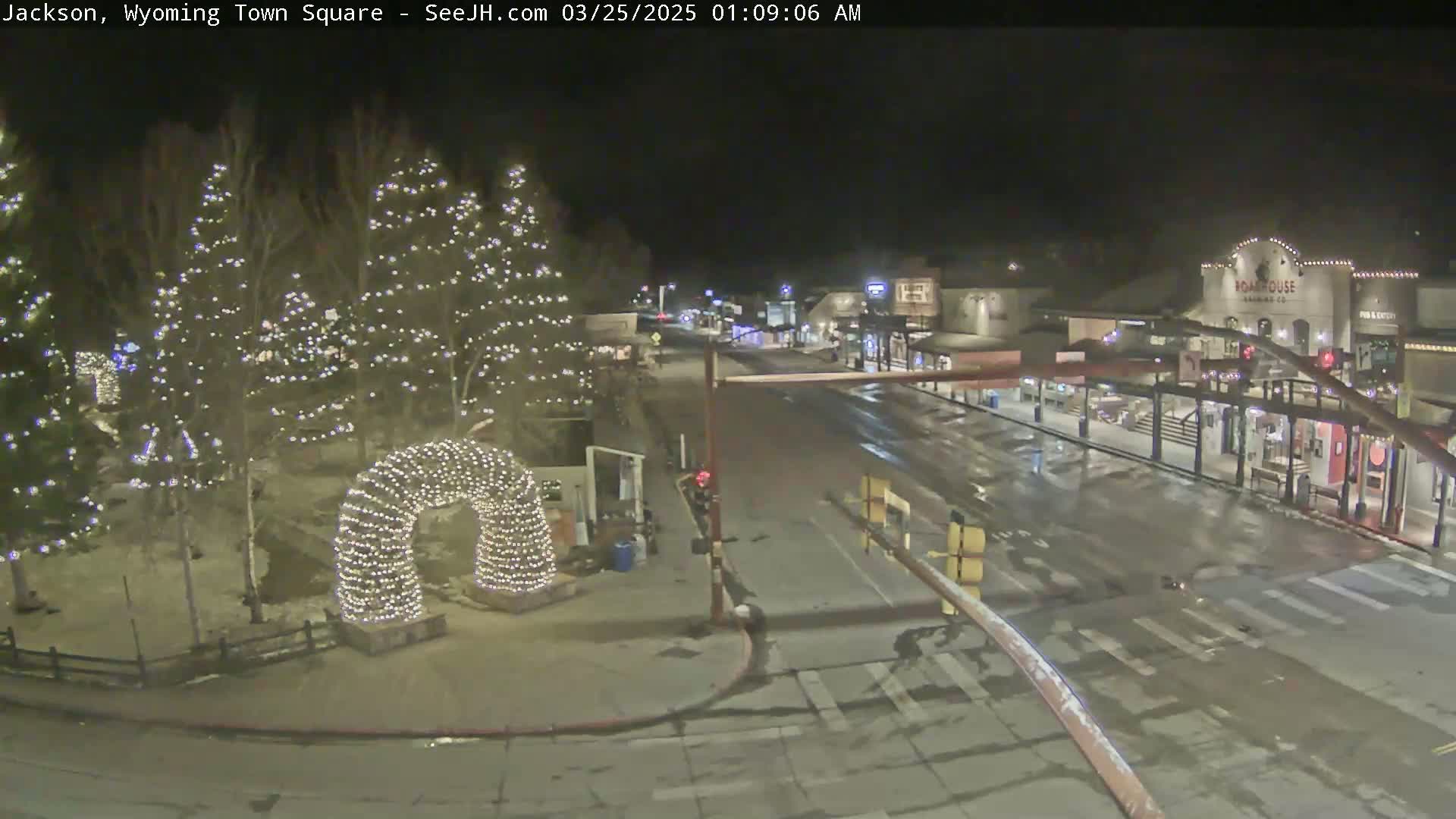 A nighttime view of a town square, lightly snow-covered, with Christmas lights illuminating trees and an archway, and wet streets showing some light traffic.