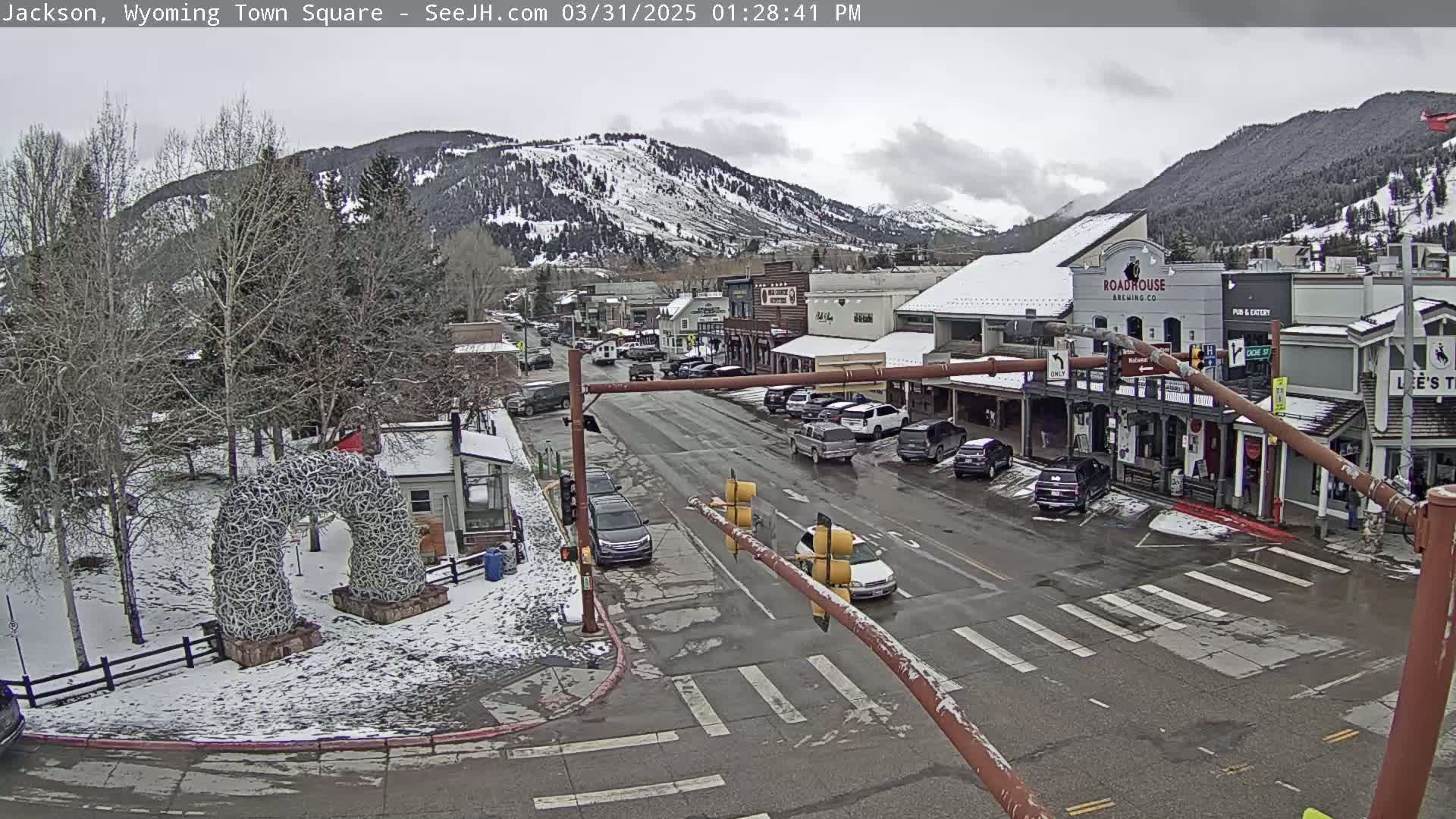 A snow-dusted town square with parked cars and buildings, situated at the base of a snow-covered mountain range under a cloudy sky.