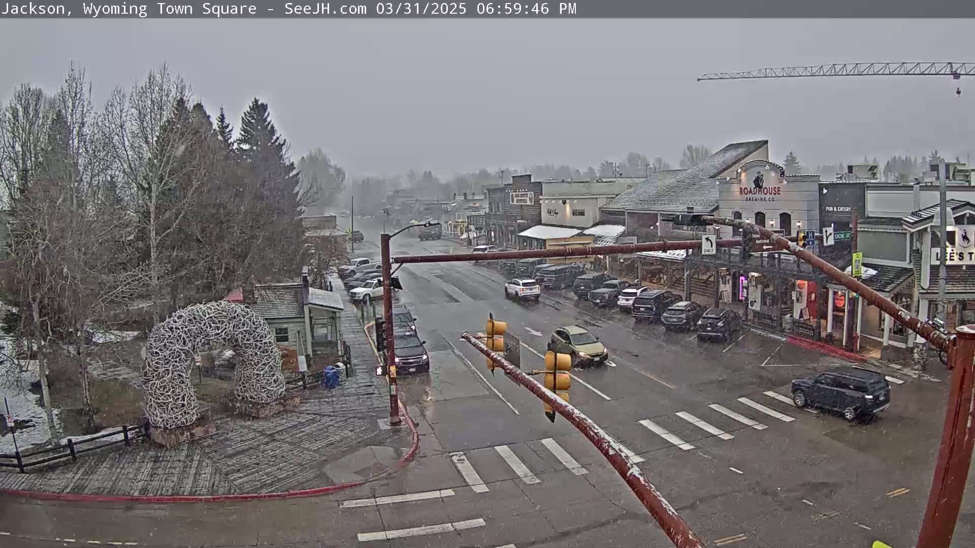 A town square with snow falling, featuring a street with parked and moving cars, buildings, and a large antler arch.