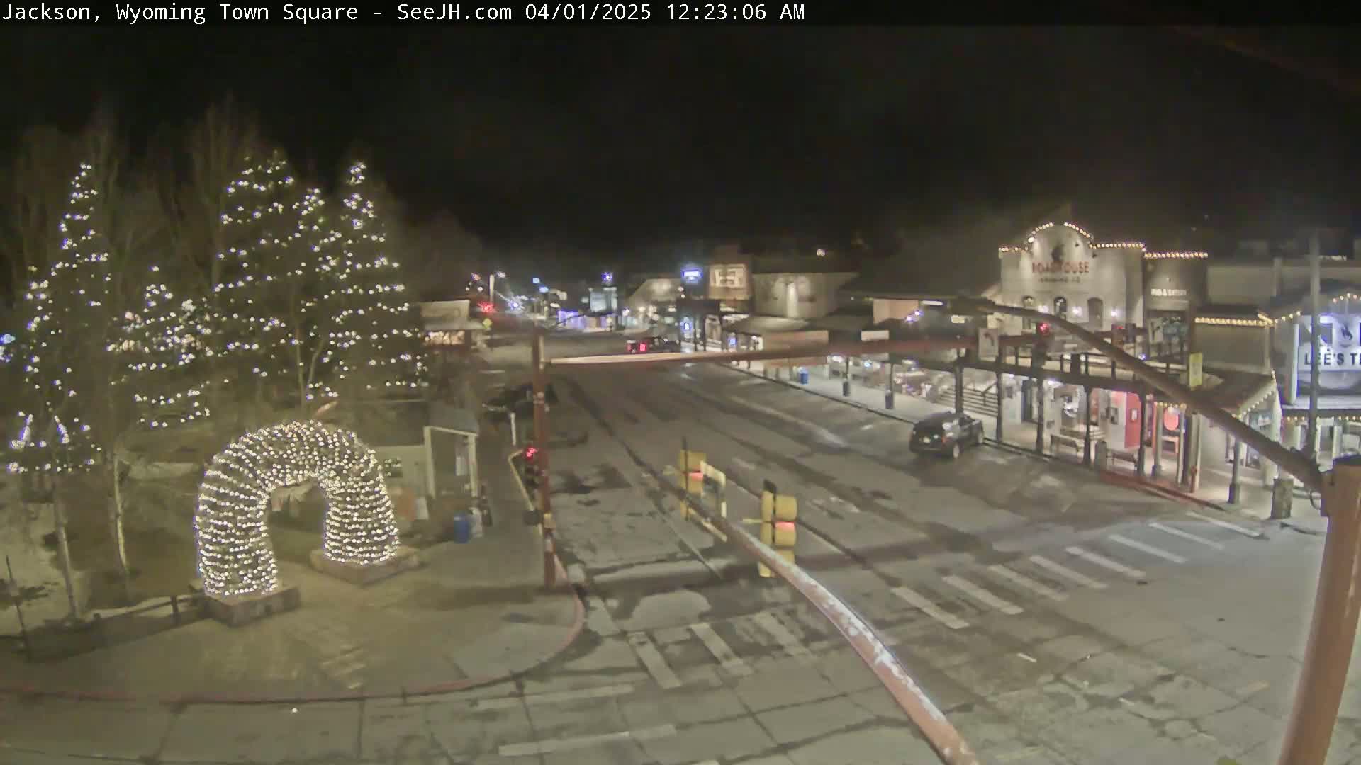 A snow-covered town square at night, illuminated by Christmas lights on trees and an archway, with buildings and vehicles visible in the distance.