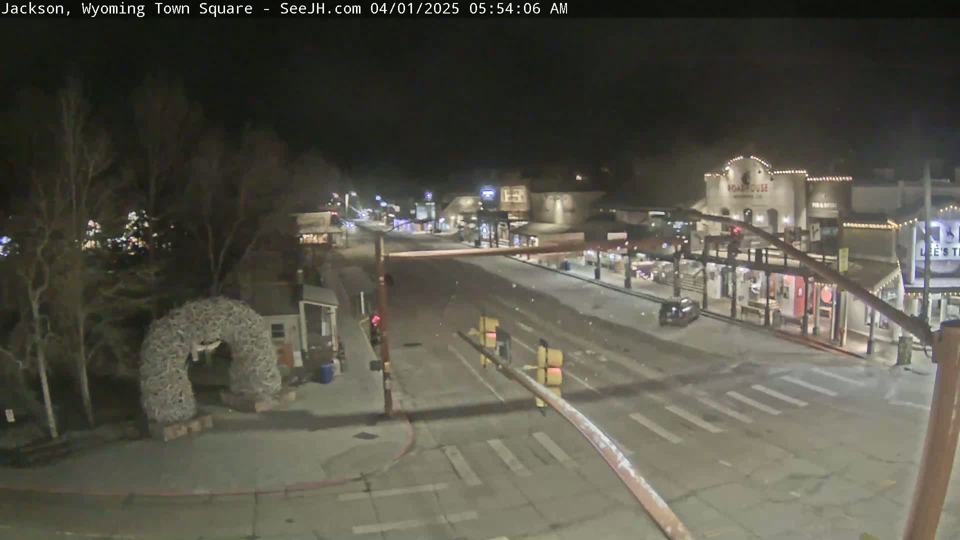 A mostly empty town square at night, lightly snow-covered, with some buildings illuminated and a large antler arch visible in the foreground.