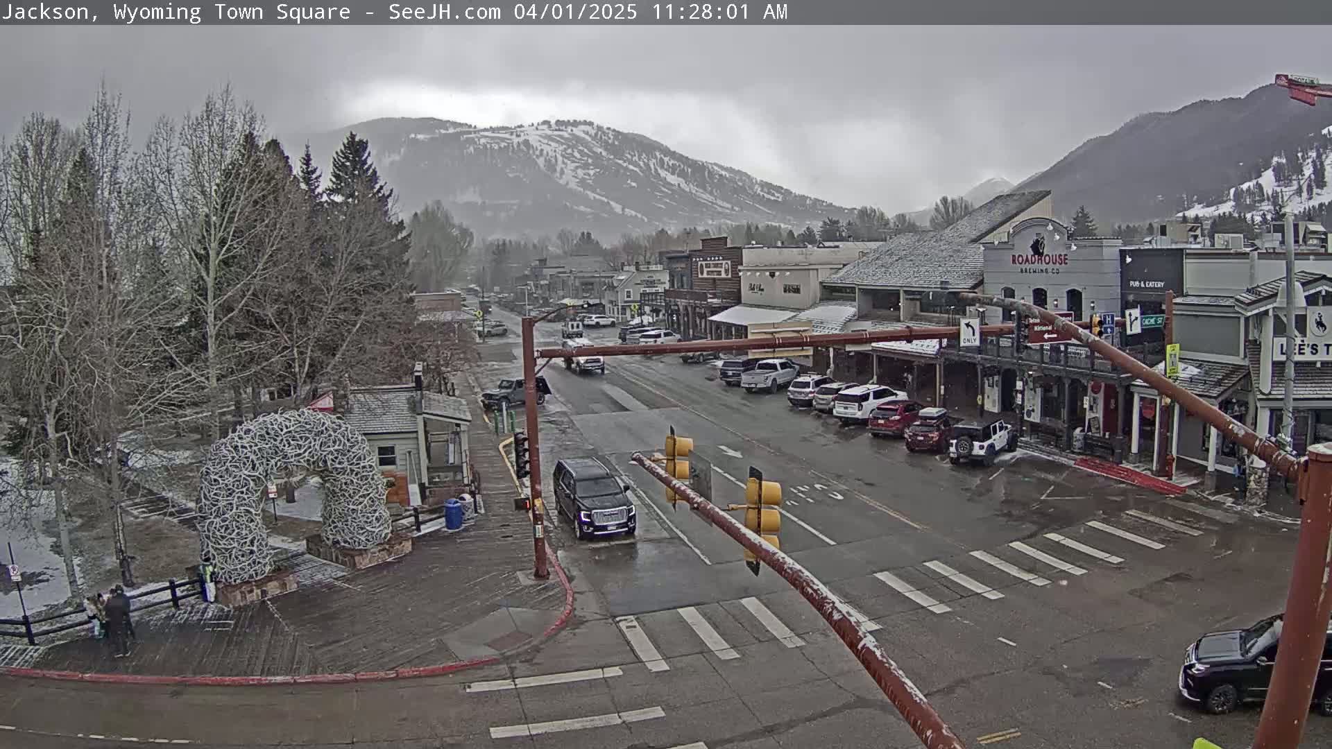 A town square with cars parked along the sides of a street, under an overcast sky, with snow-capped mountains in the background.