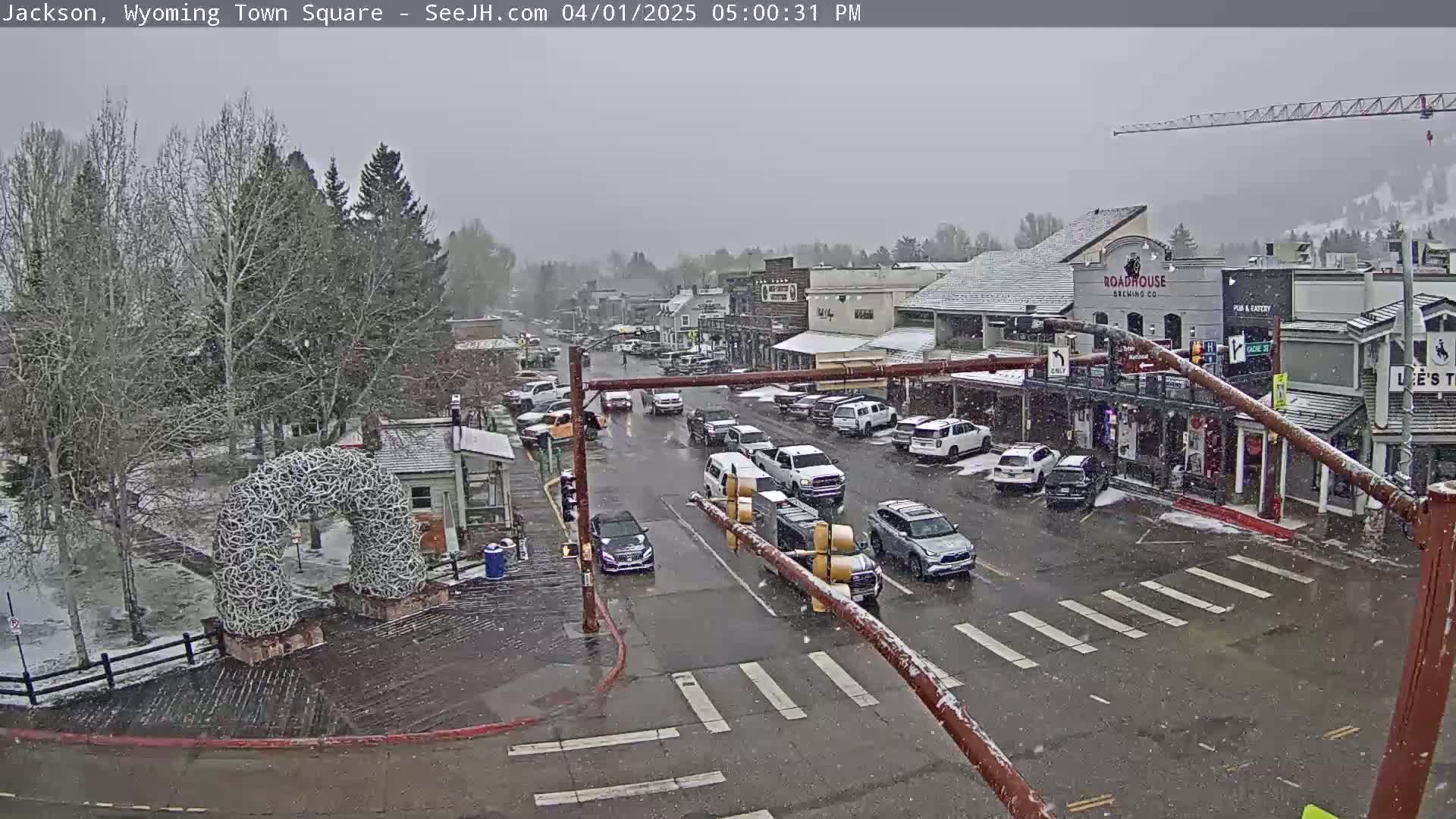 A snow-covered town square with cars parked along the sides of a street, and several vehicles traveling on the street.
