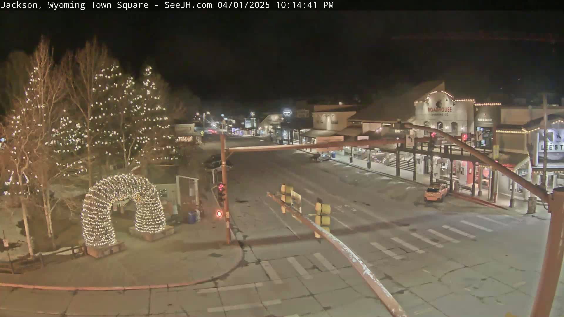 A nighttime view of a town square, featuring a light-covered archway and trees illuminated with lights, with a lightly snow-covered street and buildings.