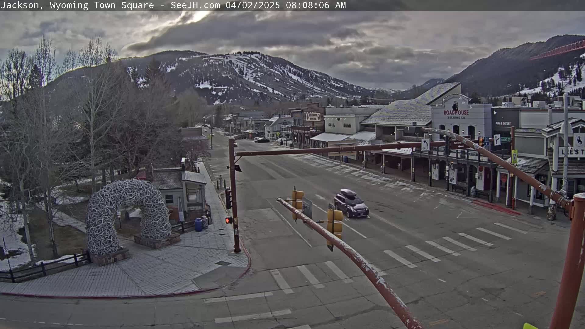 A mostly cloudy morning reveals a snow-dusted town square with a few cars driving down the street, nestled between snow-capped mountains and buildings.