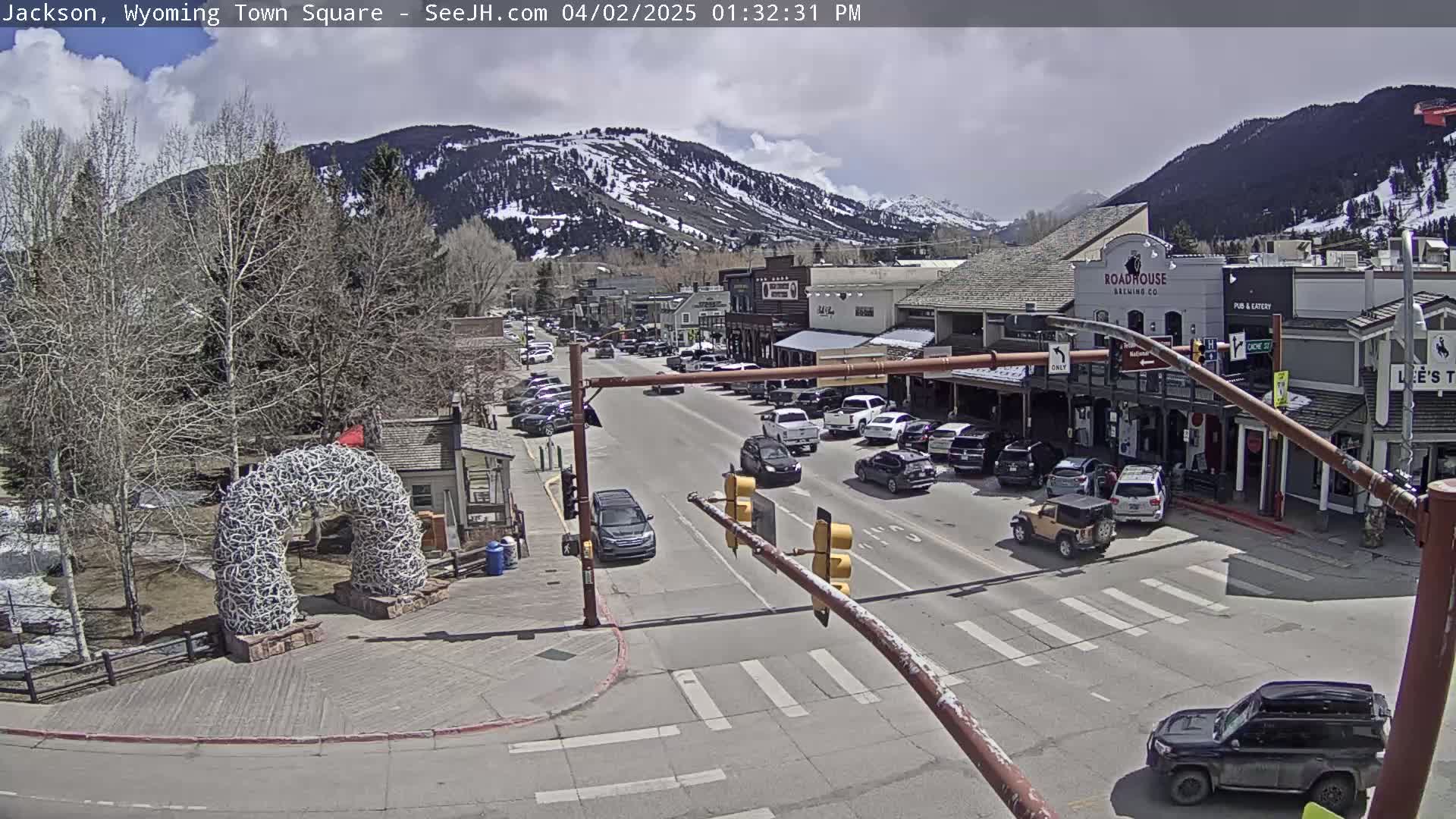 A partly cloudy day reveals a town square with a street scene and parked cars, nestled at the base of snow-capped mountains.