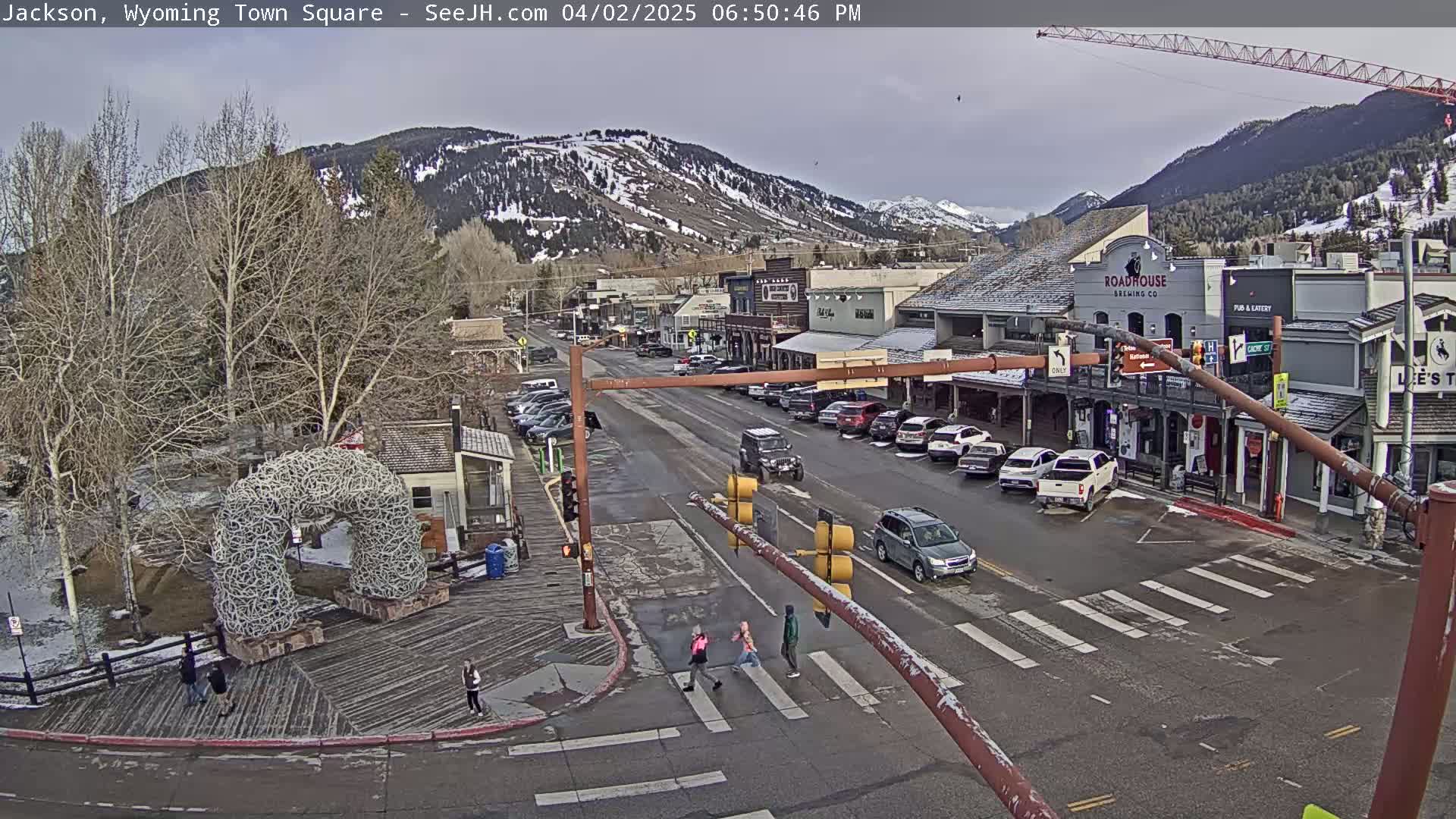 A town square scene shows a street with parked and moving cars, pedestrians crossing at a crosswalk, and buildings under a mostly cloudy sky, with snow-capped mountains in the background.
