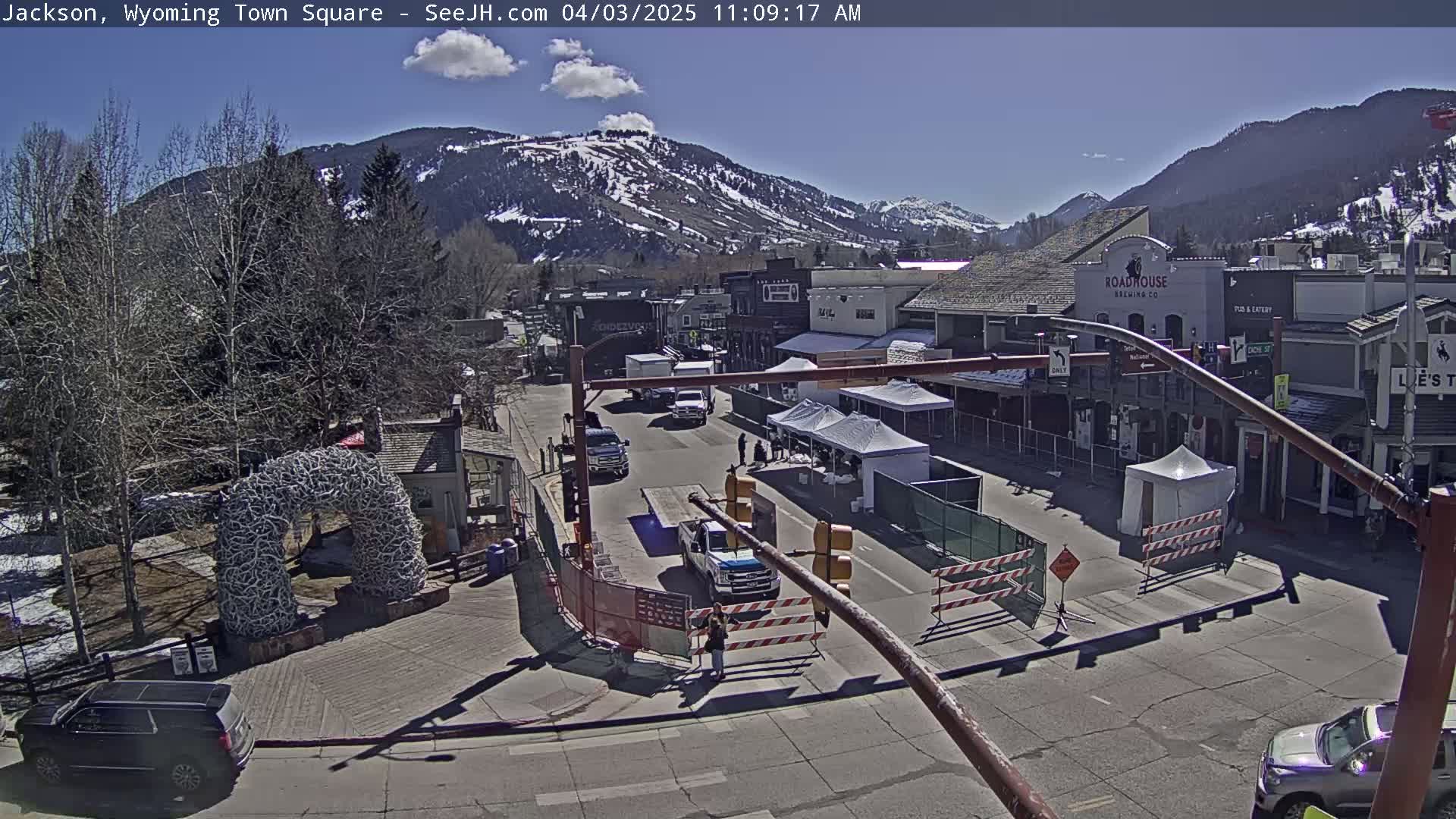 A partly sunny day shows a town square with buildings, vehicles, and temporary structures, set against a backdrop of snow-capped mountains.